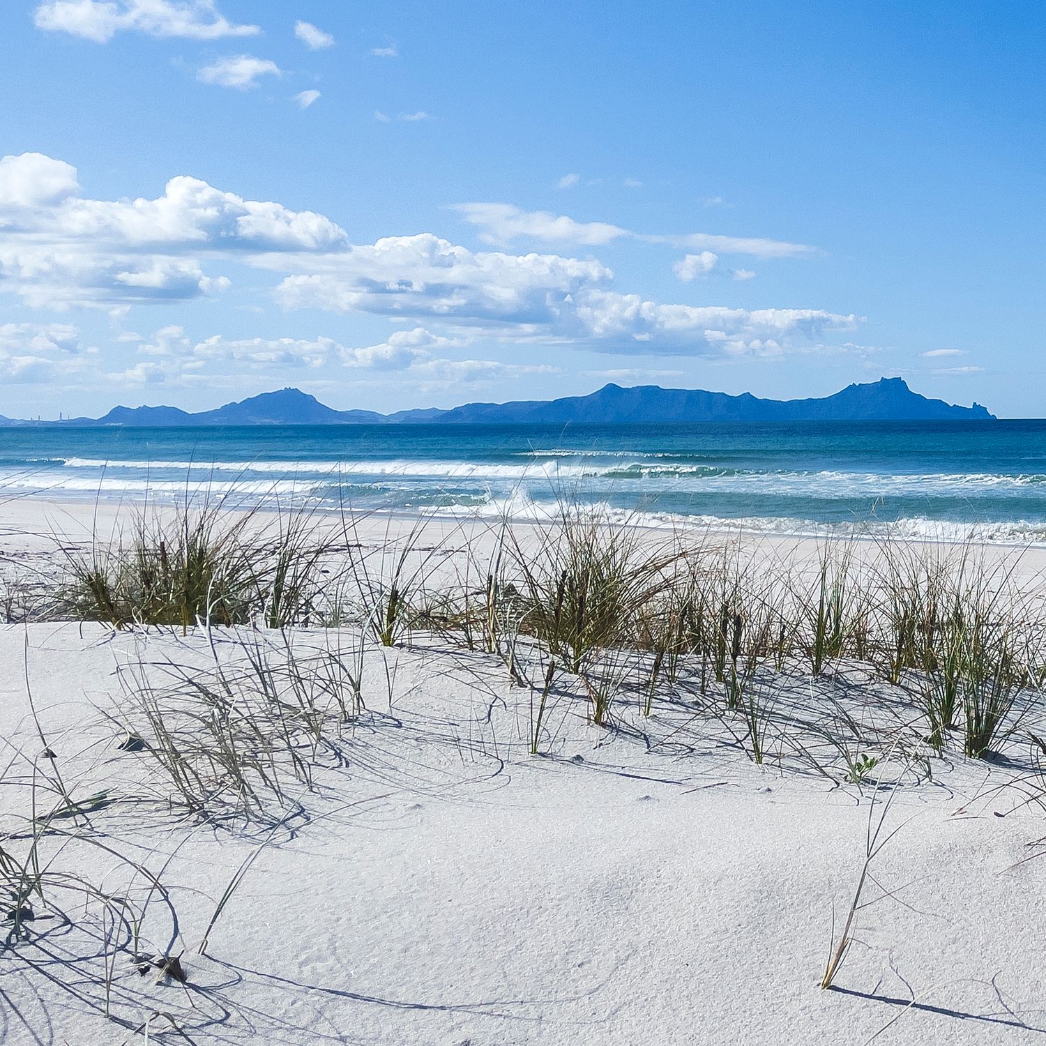 White sand dunes with coastal grass, turquoise ocean waves, and mountain range under blue sky at a pristine beach.