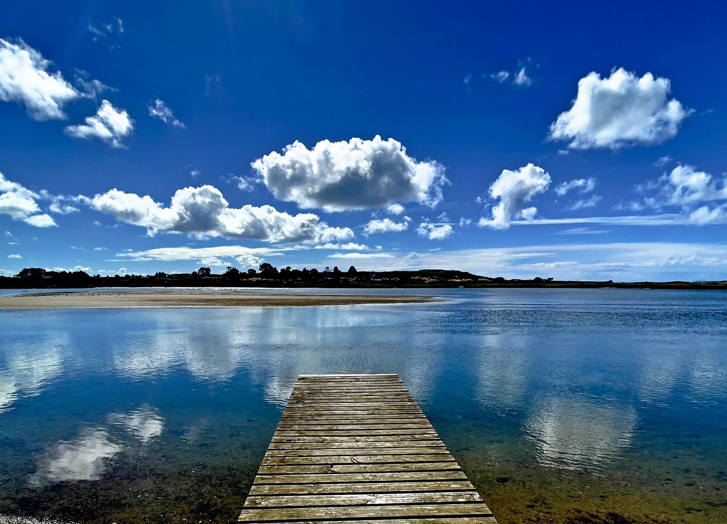 Wooden dock extending into calm reflective lake under vivid blue sky with white clouds mirrored on water surface.
