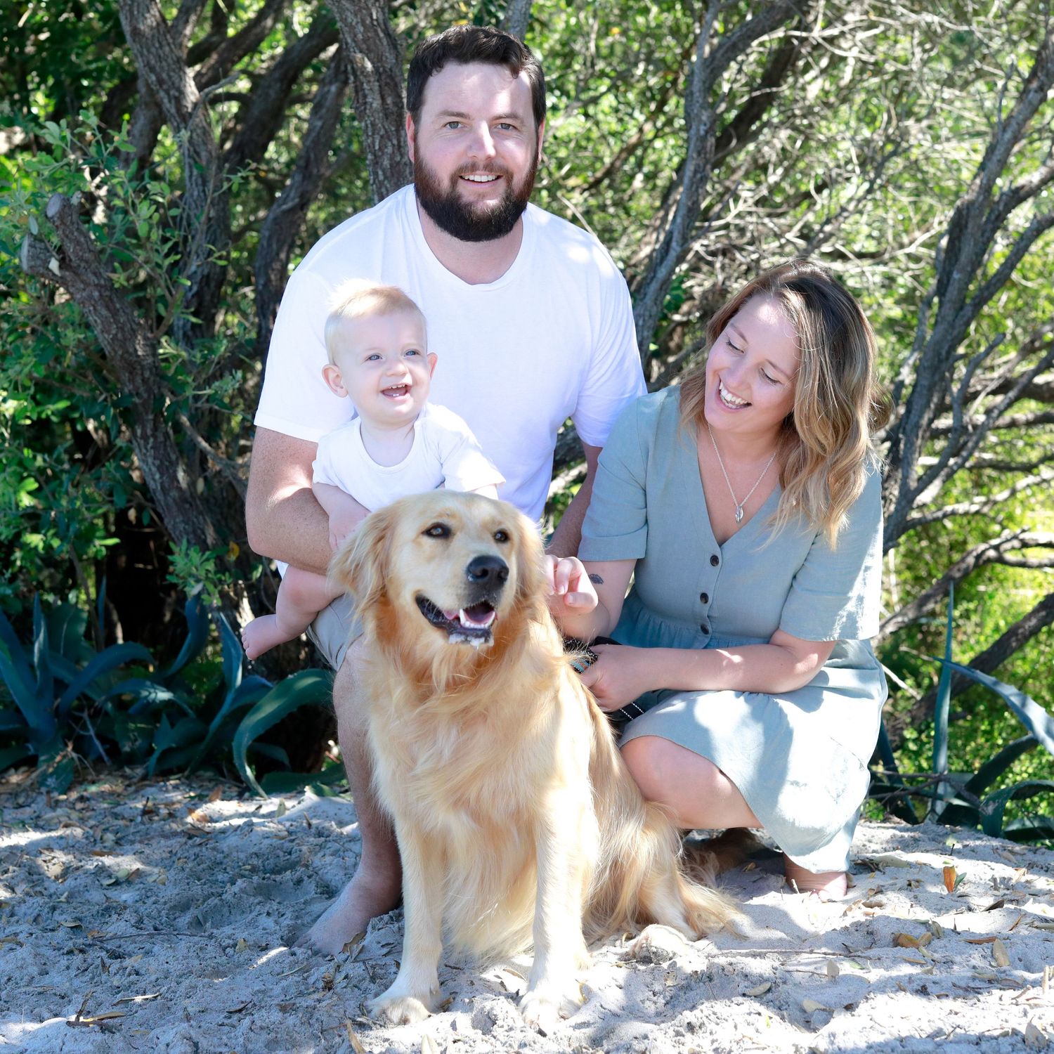 A happy family with a man, woman, baby, and golden retriever dog posing together on a sandy beach.
