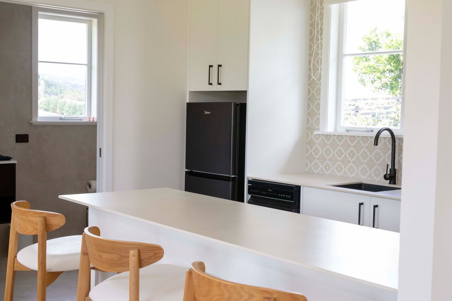 Modern white kitchen with bar seating, black appliances, patterned backsplash, and natural wood stools.