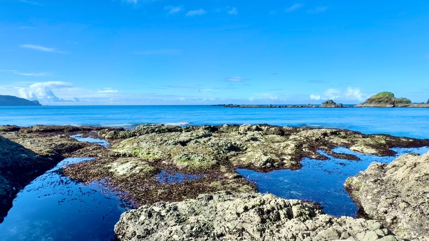 Rocky tide pools along a tropical coastline with calm blue ocean waters and small islands visible on the horizon.