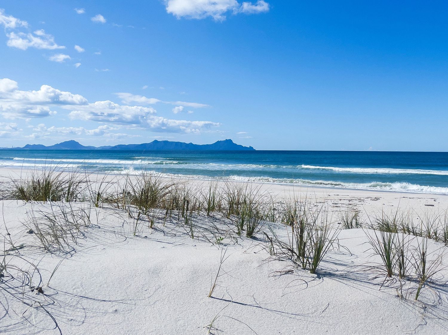 White sand dunes with coastal grass overlook a pristine turquoise beach under a bright blue sky with distant mountains.