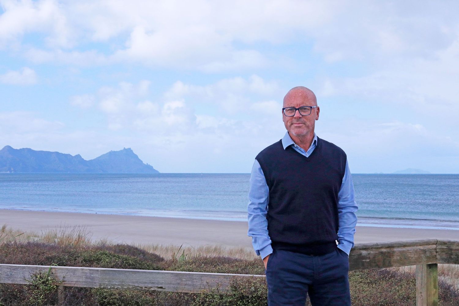 Man standing on a scenic beach with mountain views and calm ocean waters in the background.