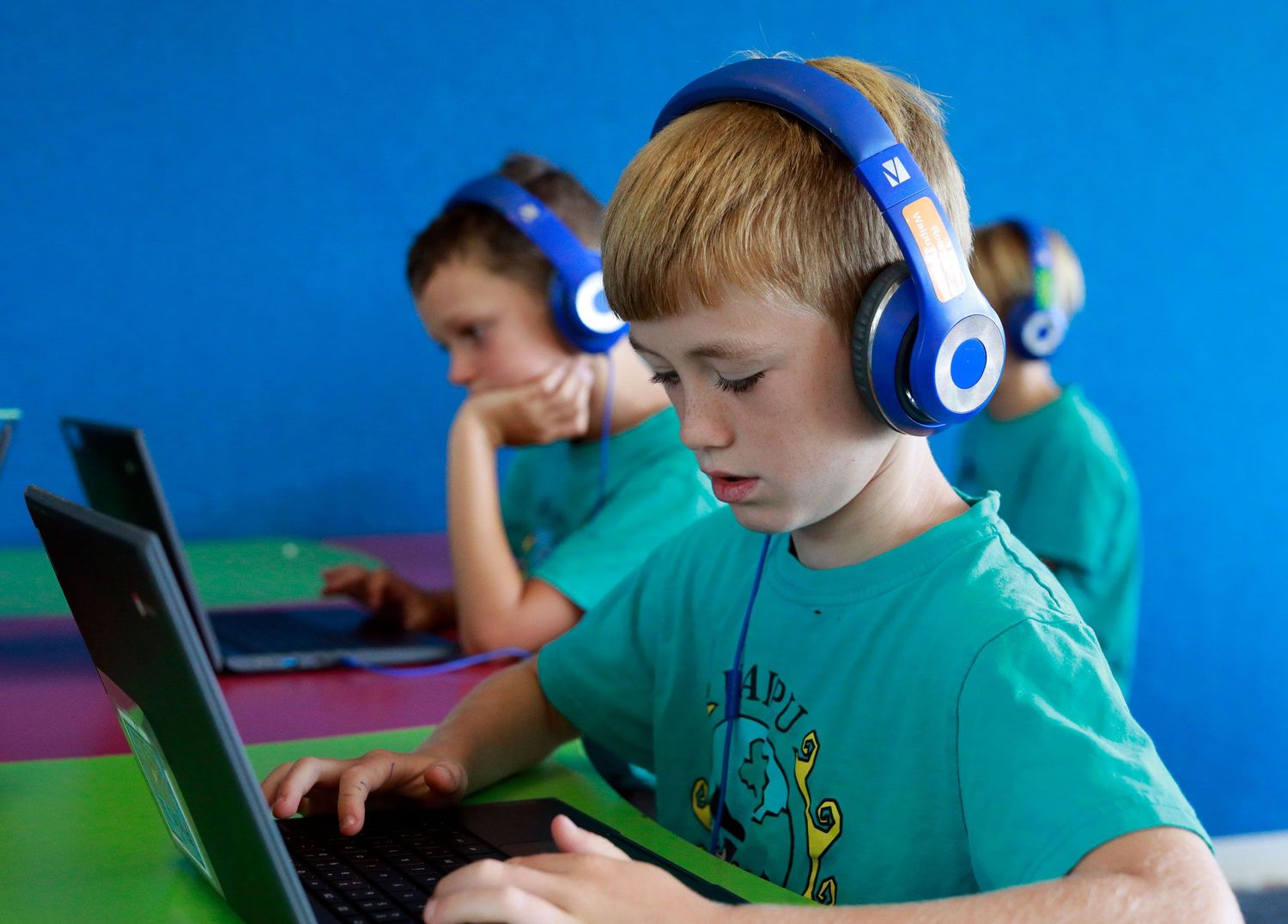Two children wearing blue headphones use laptops in a classroom with colorful walls.