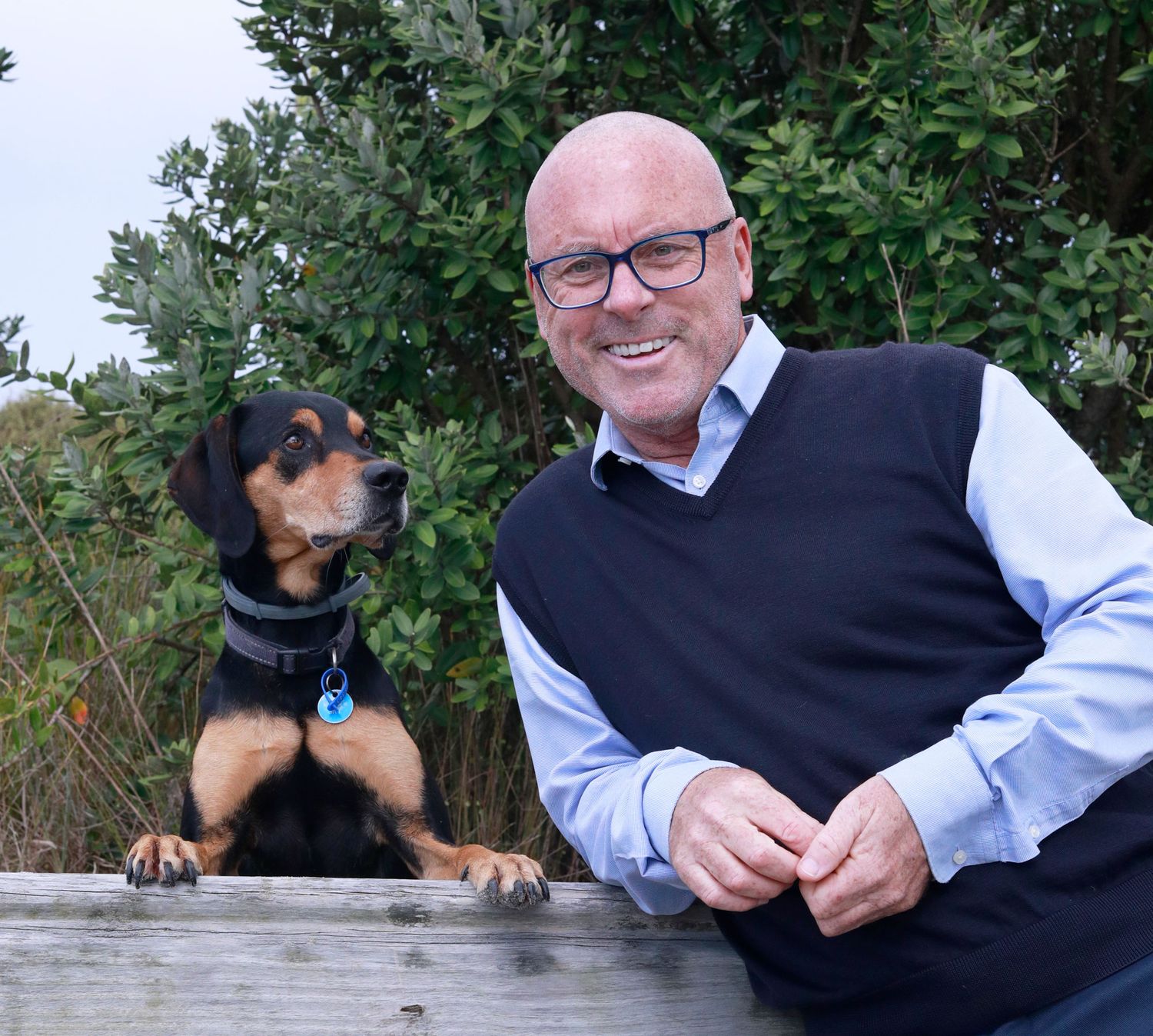 Smiling bald man with glasses in a black vest poses with a black and tan puppy on a wooden fence outdoors.