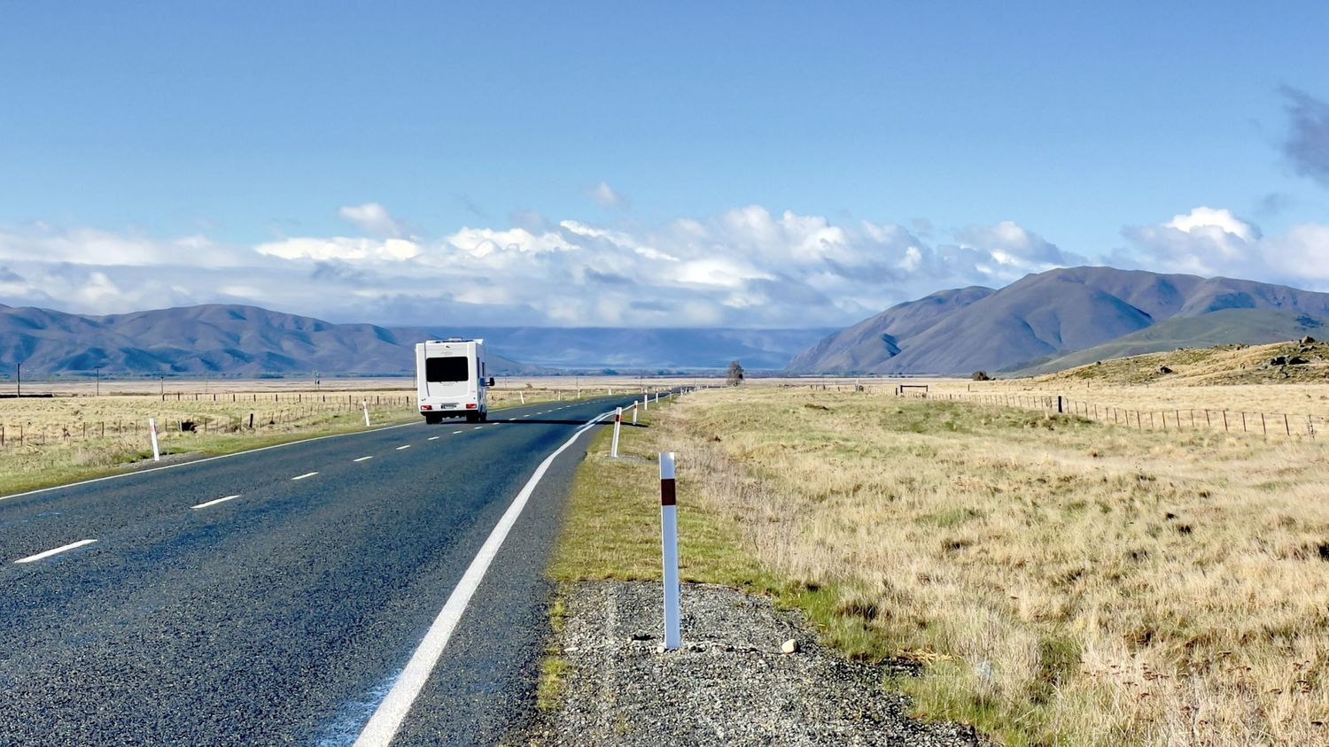 A white campervan travels along a straight rural highway through golden grasslands with mountain ranges under blue skies.