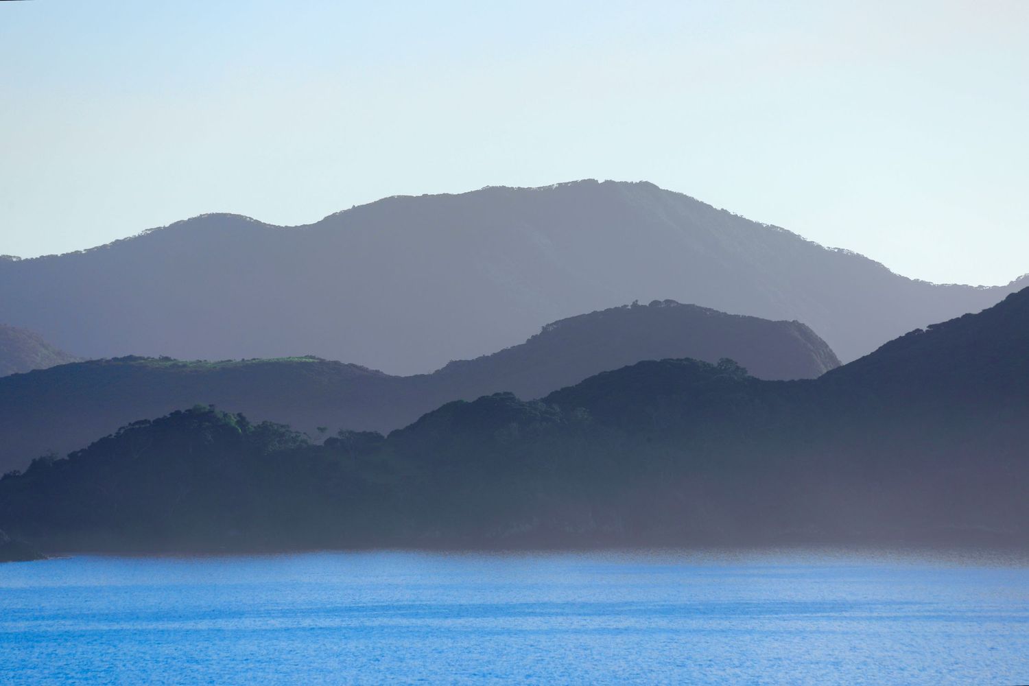 Misty blue mountains layered in haze rise above a calm blue Taupiri Bay in a serene coastal landscape.