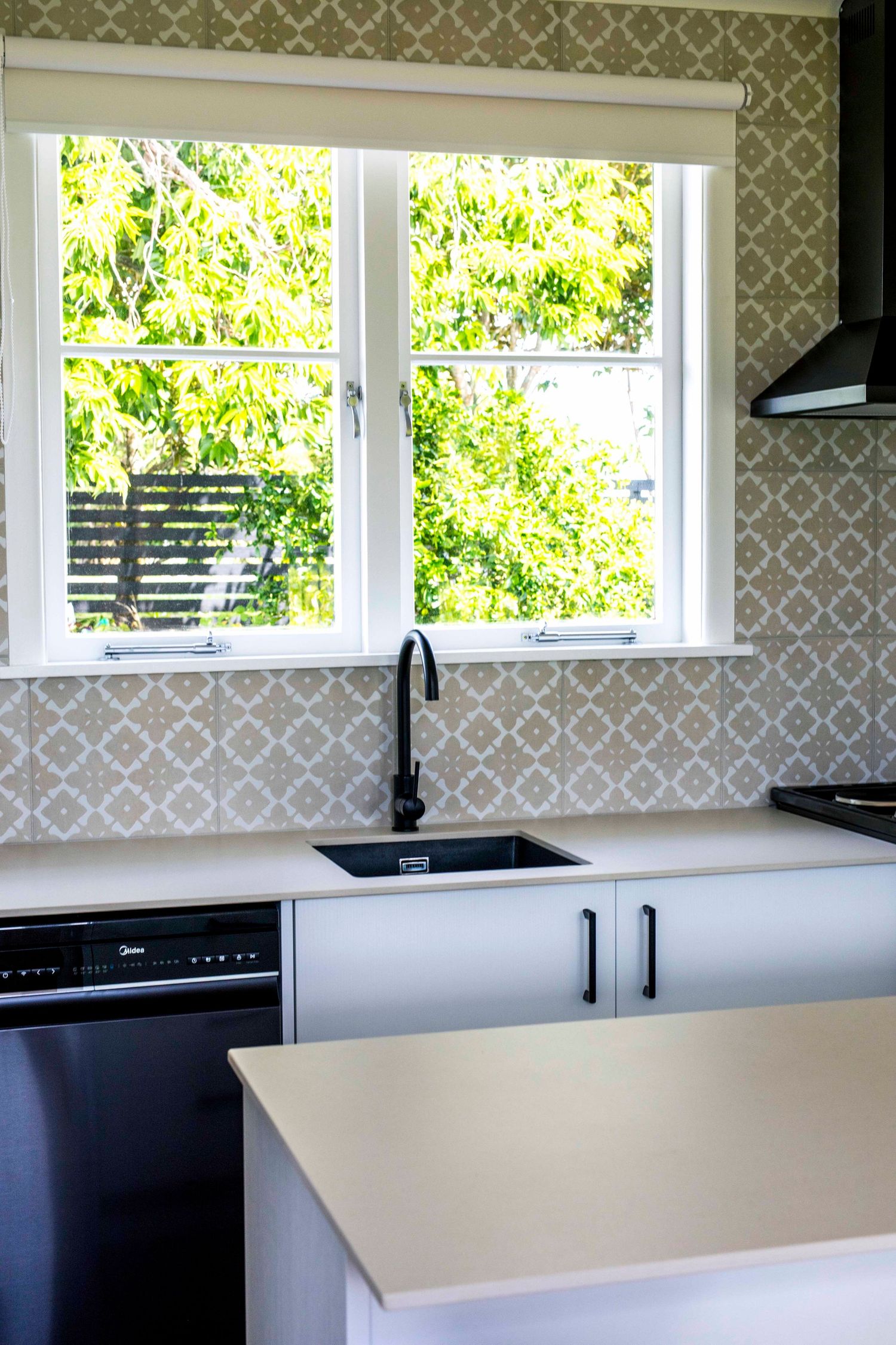 Modern white kitchen with black sink faucet, patterned tile backsplash, and bright window overlooking lush green trees.