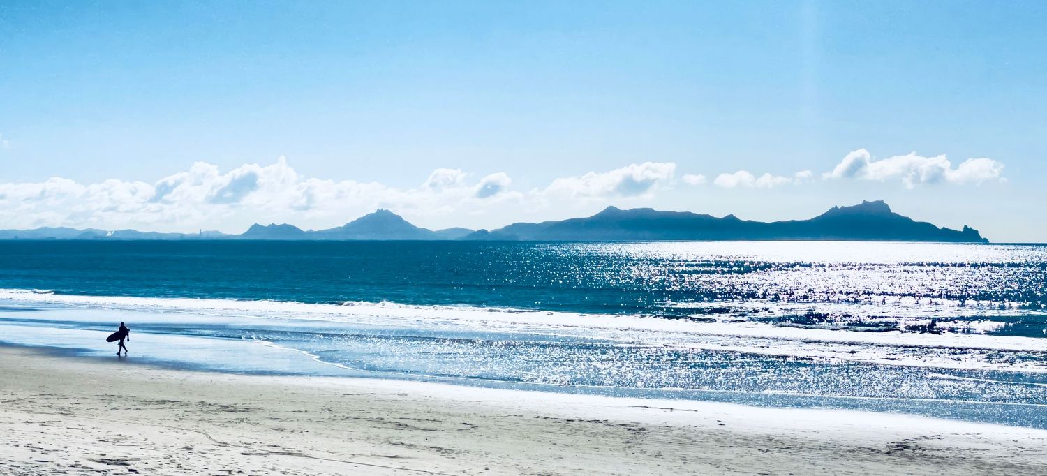 Surfer walks along a serene beach with sparkling ocean waters and distant island mountains under a bright blue sky.