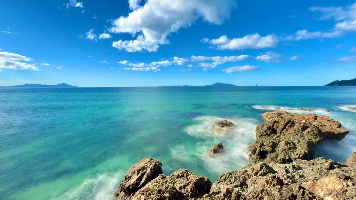 Rocky coastline with turquoise ocean waters under a bright blue sky with white clouds and distant mountains.