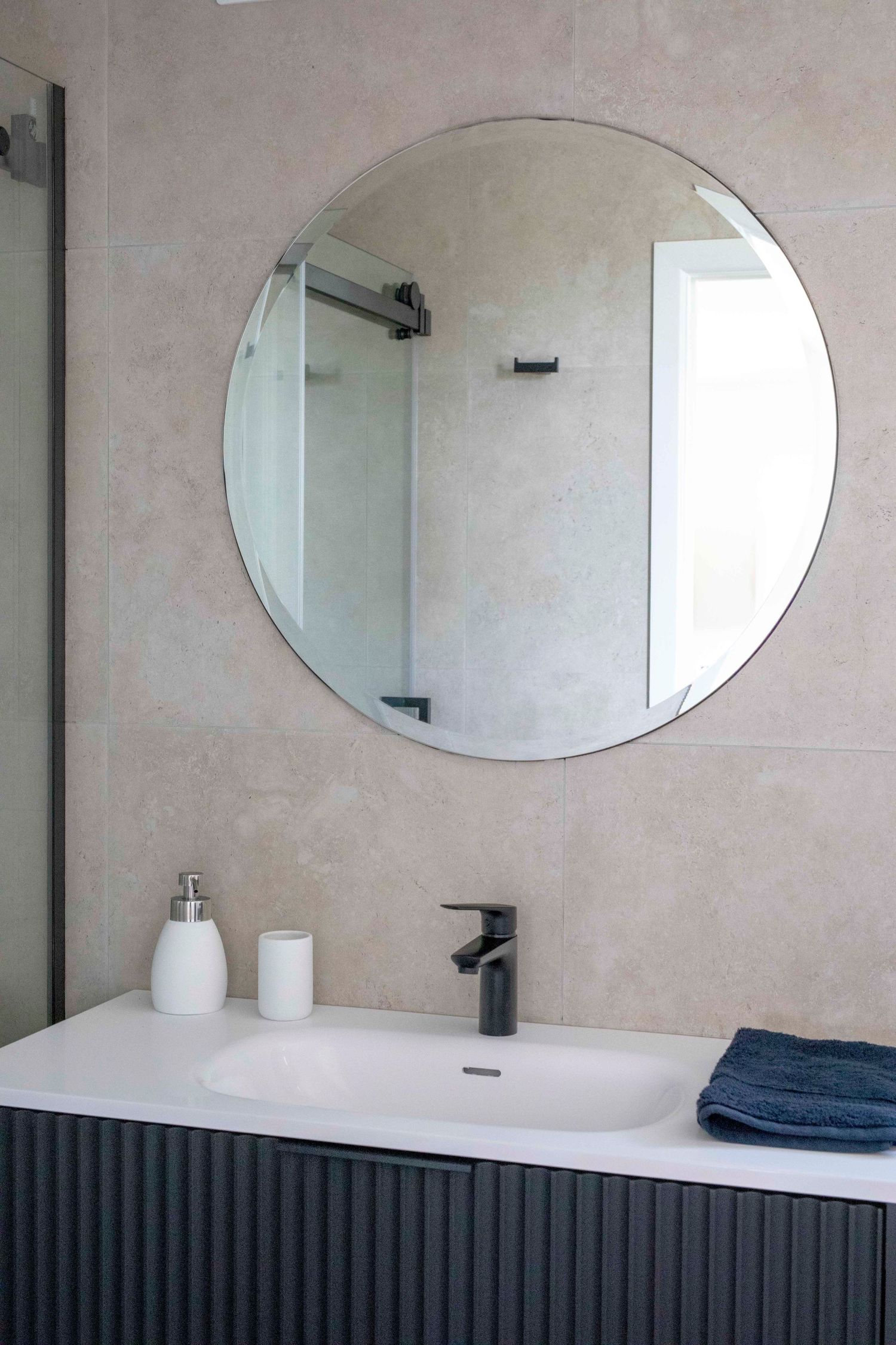 Modern bathroom vanity with round mirror, black faucet, white sink, soap dispenser, and folded blue towel on dark cabinet.