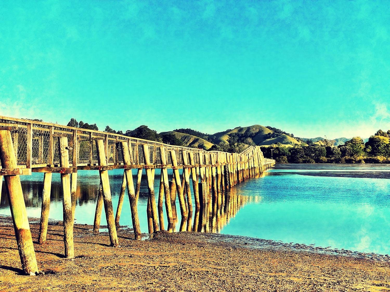 Wooden pier stretching over calm blue water with sandy shore and green hills in background under clear turquoise sky.