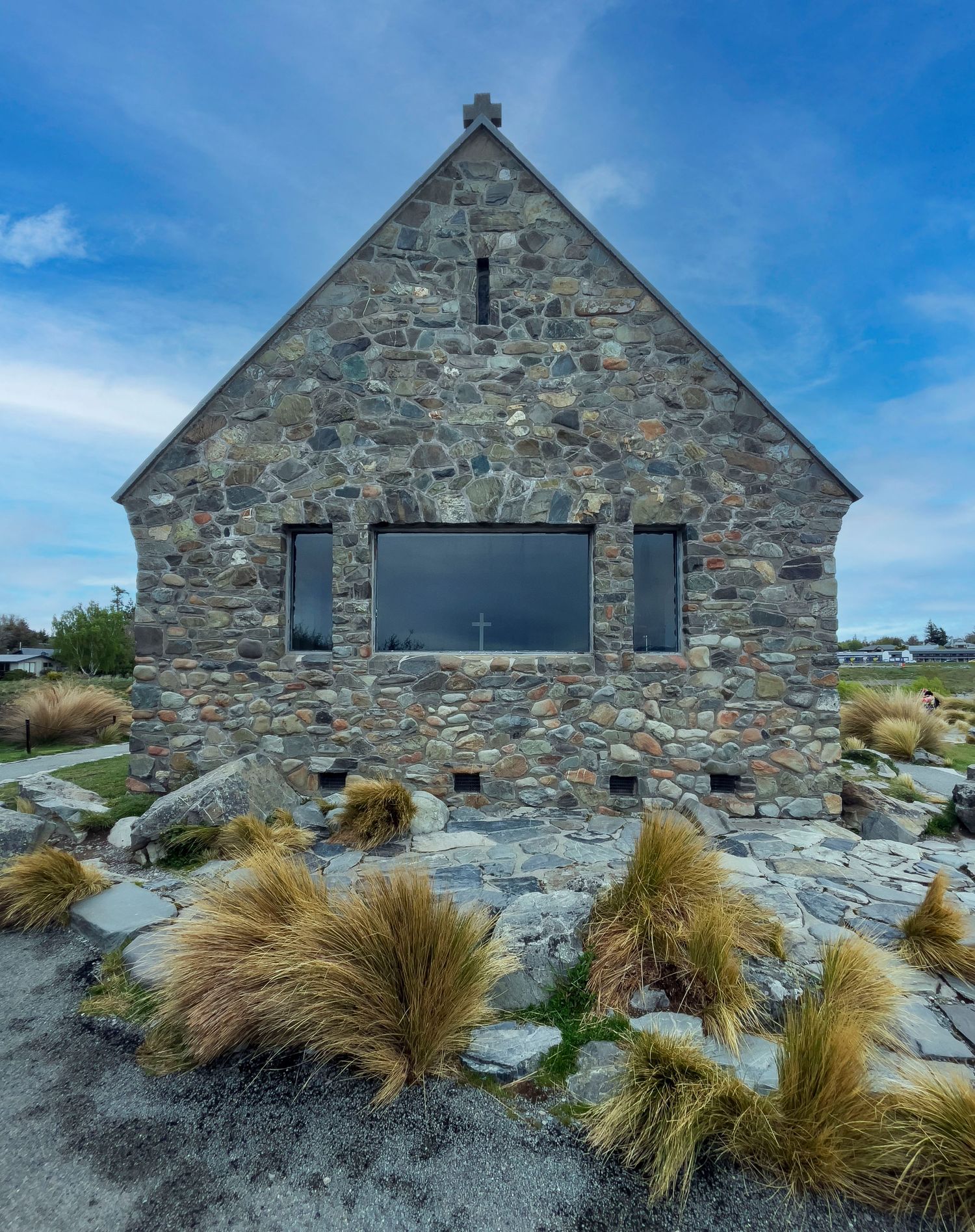 Stone church with gabled roof, large windows, and tussock grass landscaping under a blue sky.