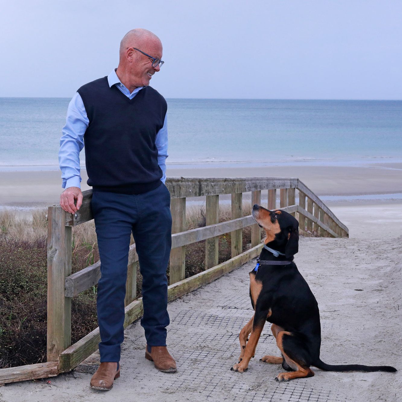 Man in navy vest and glasses stands with a black and tan dog on a beachside boardwalk path.