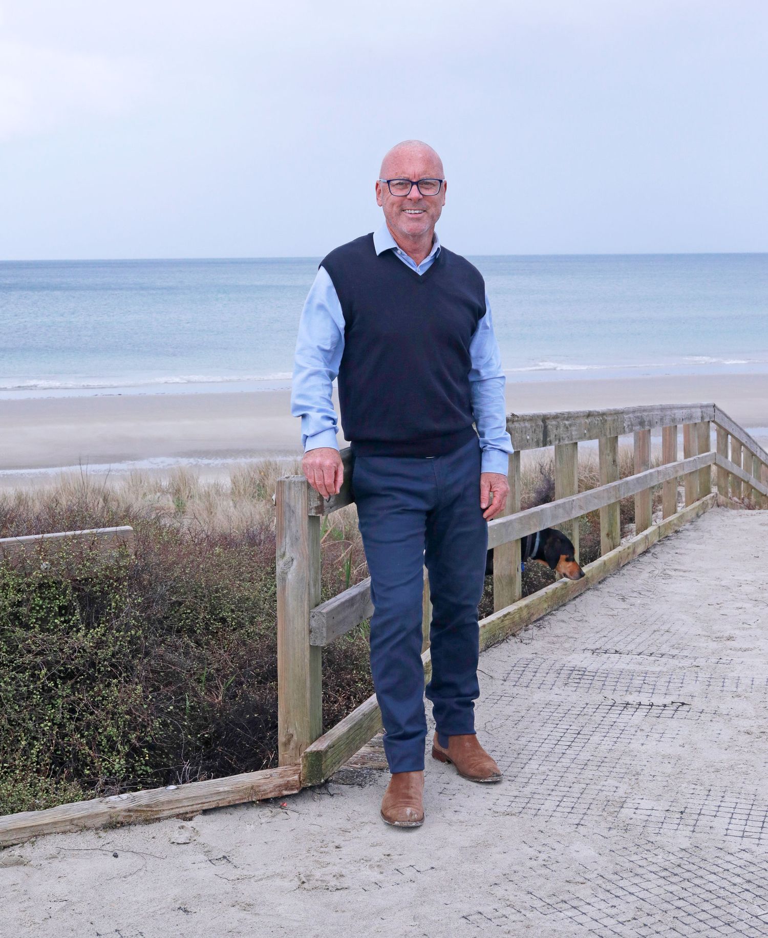 Man in navy vest and blue shirt standing on beach boardwalk with ocean view on cloudy day.