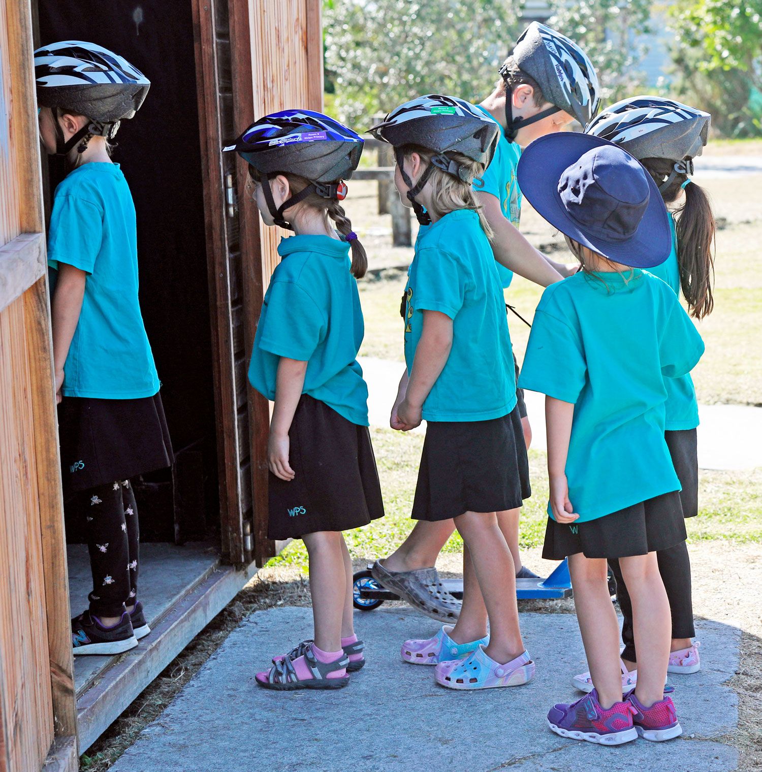 Young girls in teal uniforms and bike helmets line up outside a wooden building during a school cycling activity.