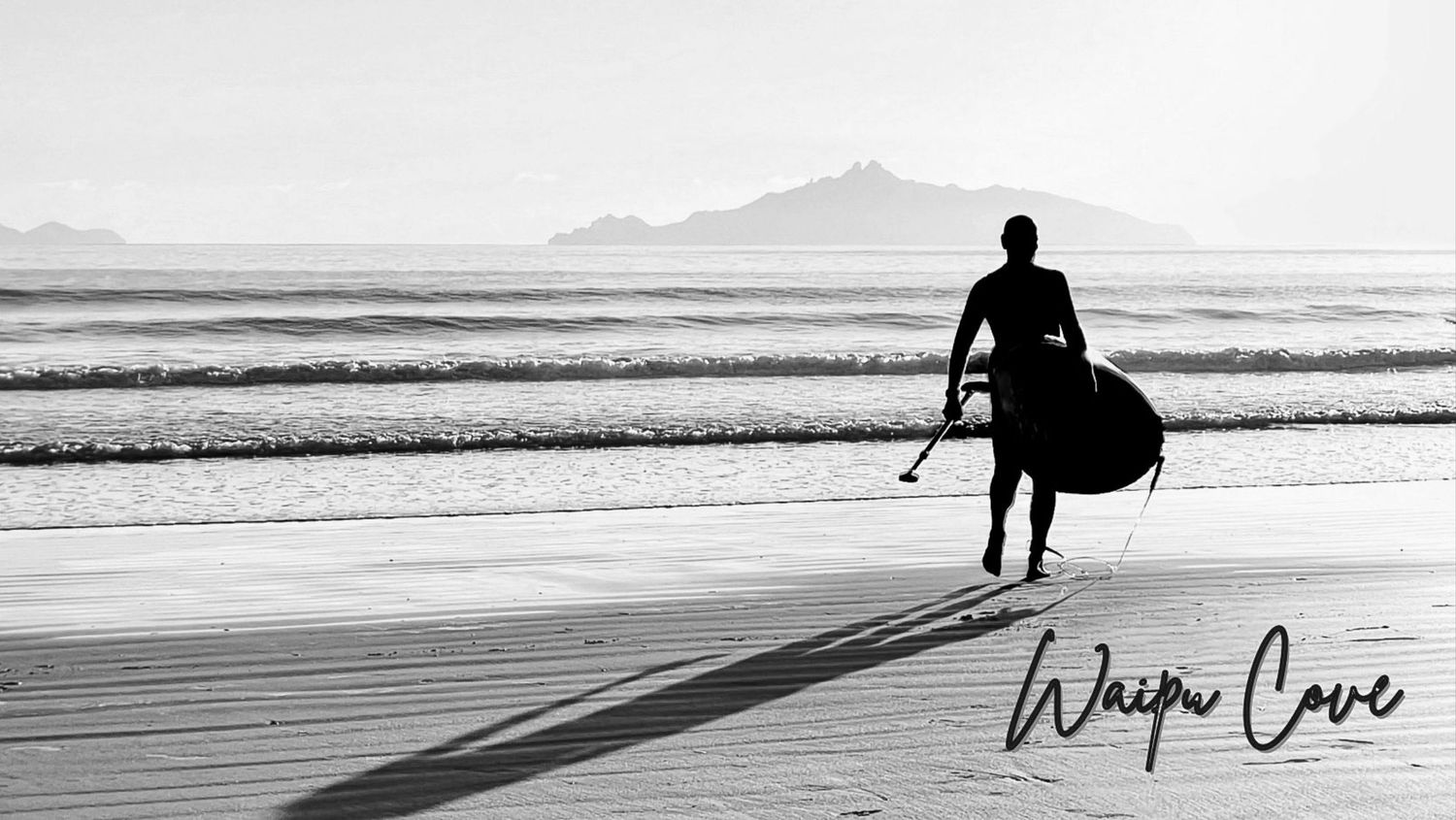 Black and white photo of a surfer carrying a board at Waipu Cove beach with mountains in the background.