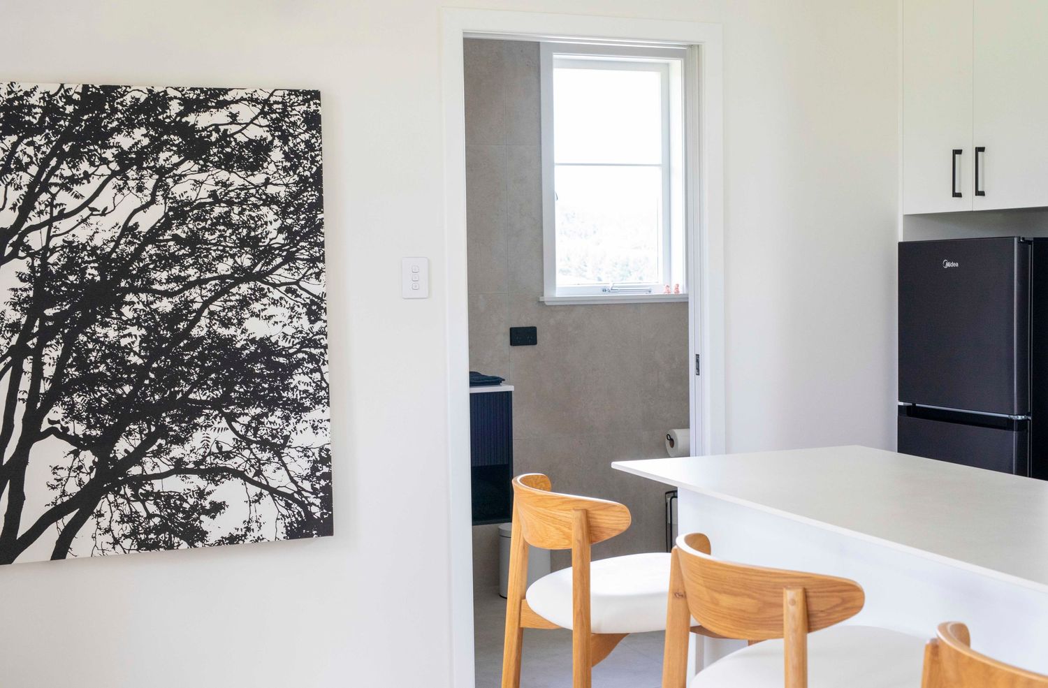 Modern minimalist dining area with wooden chairs, white table, black tree artwork, and a view into a bright hallway.