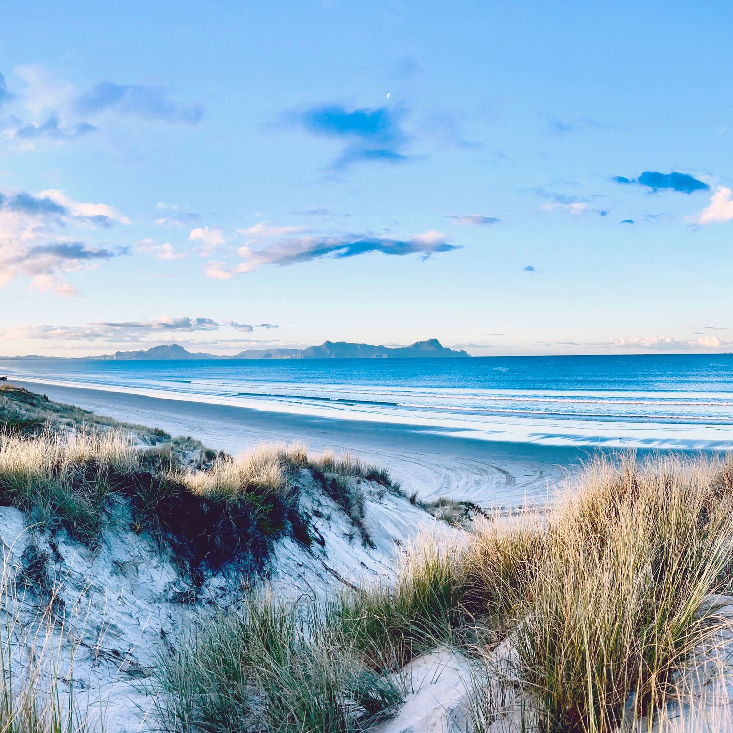 Sandy beach dunes with marram grass, turquoise ocean waves, and distant mountains under a blue sky with soft clouds.