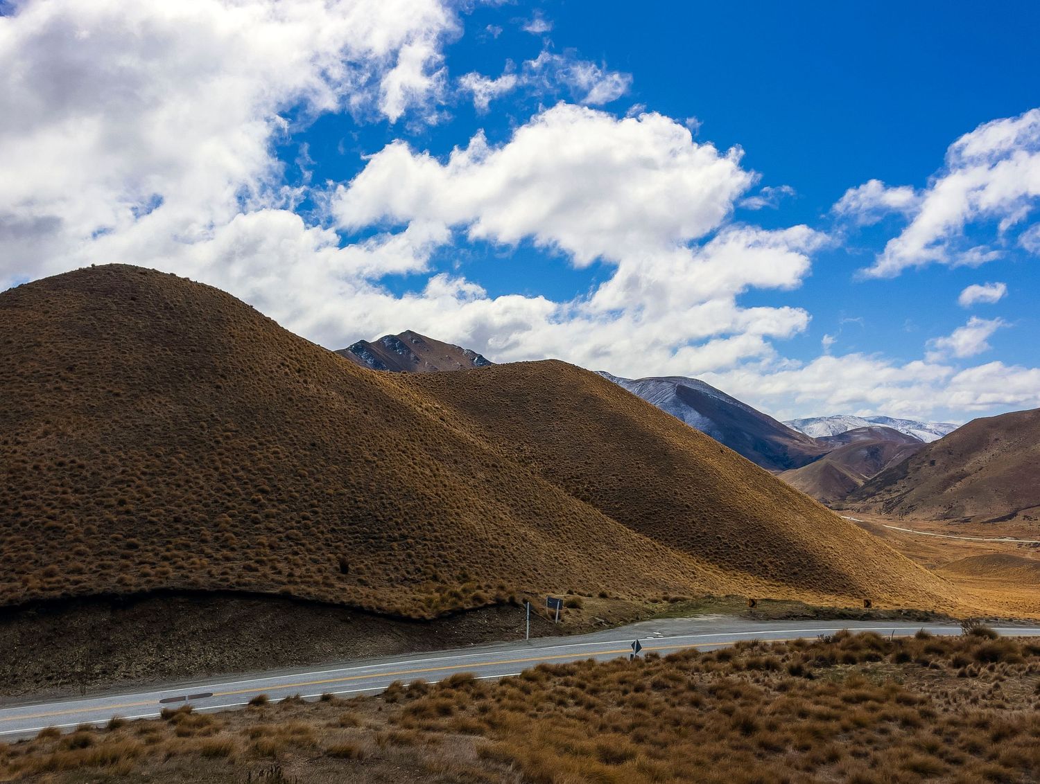 Scenic mountain road winding through golden brown hills under a vivid blue sky with white clouds in a remote highland landscape.