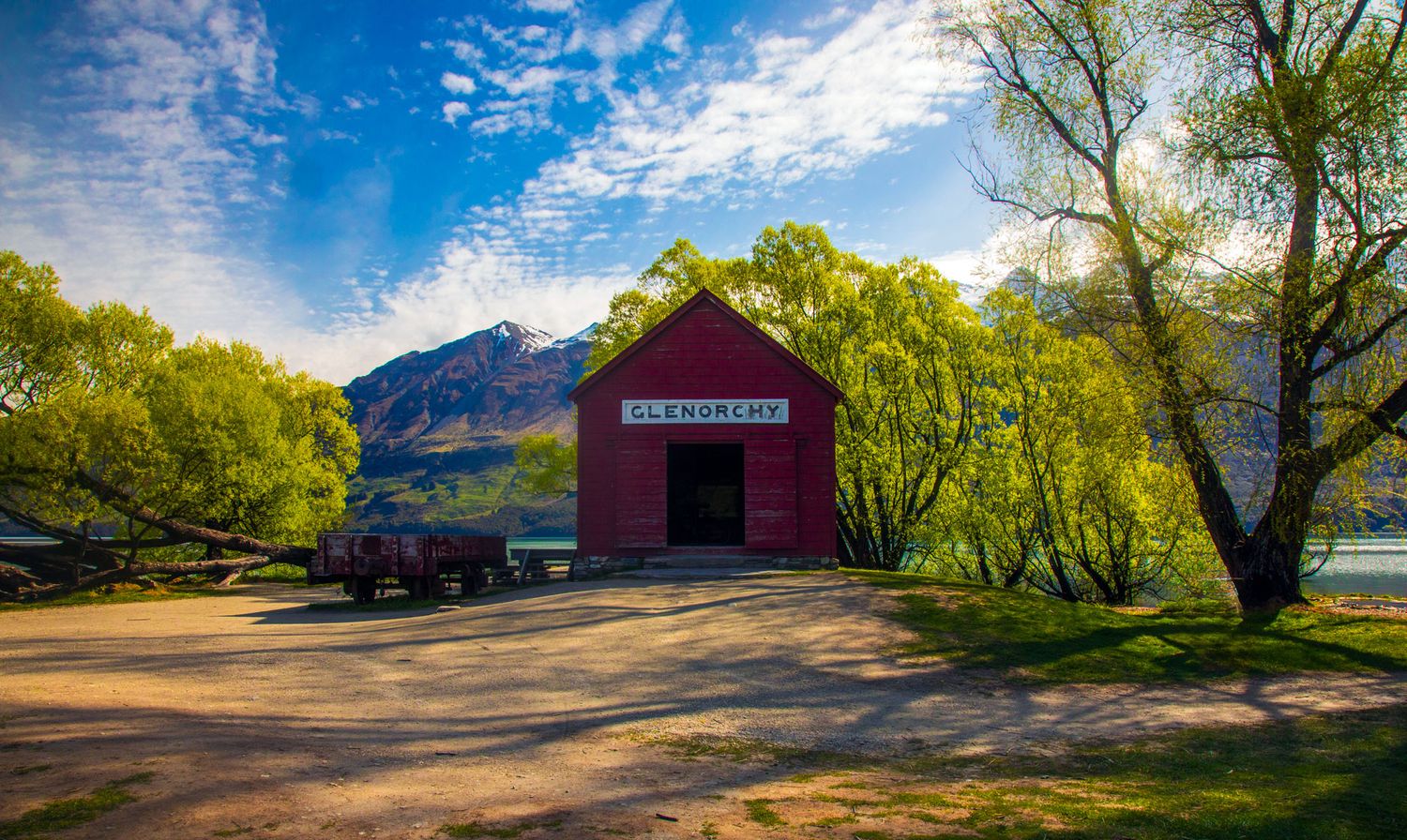Red Glenorchy boatshed beside a lake with lush green willow trees, mountains, and a blue sky in New Zealand.