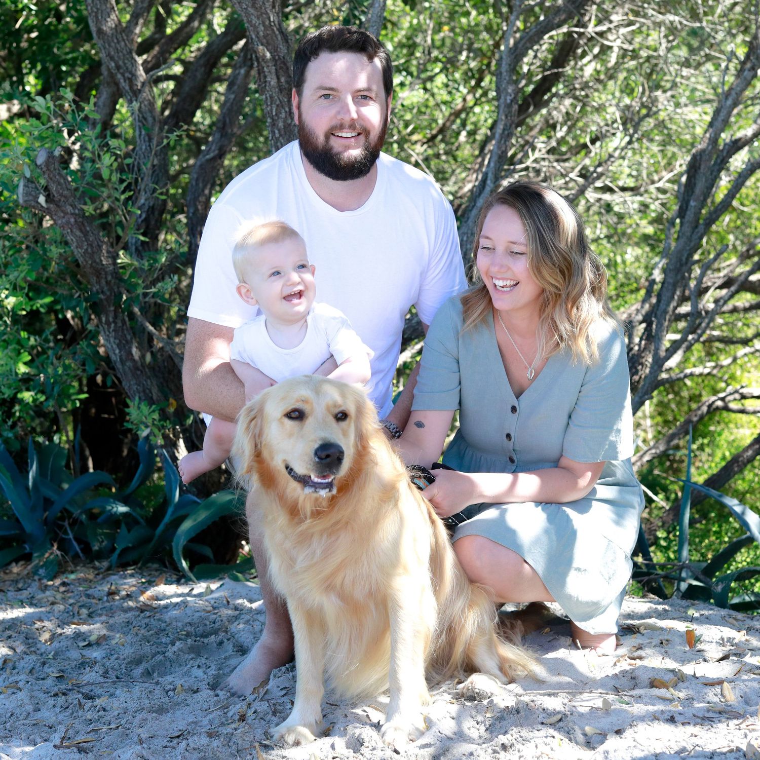 A smiling family with a baby and golden retriever dog pose together on sandy beach surrounded by lush green trees.
