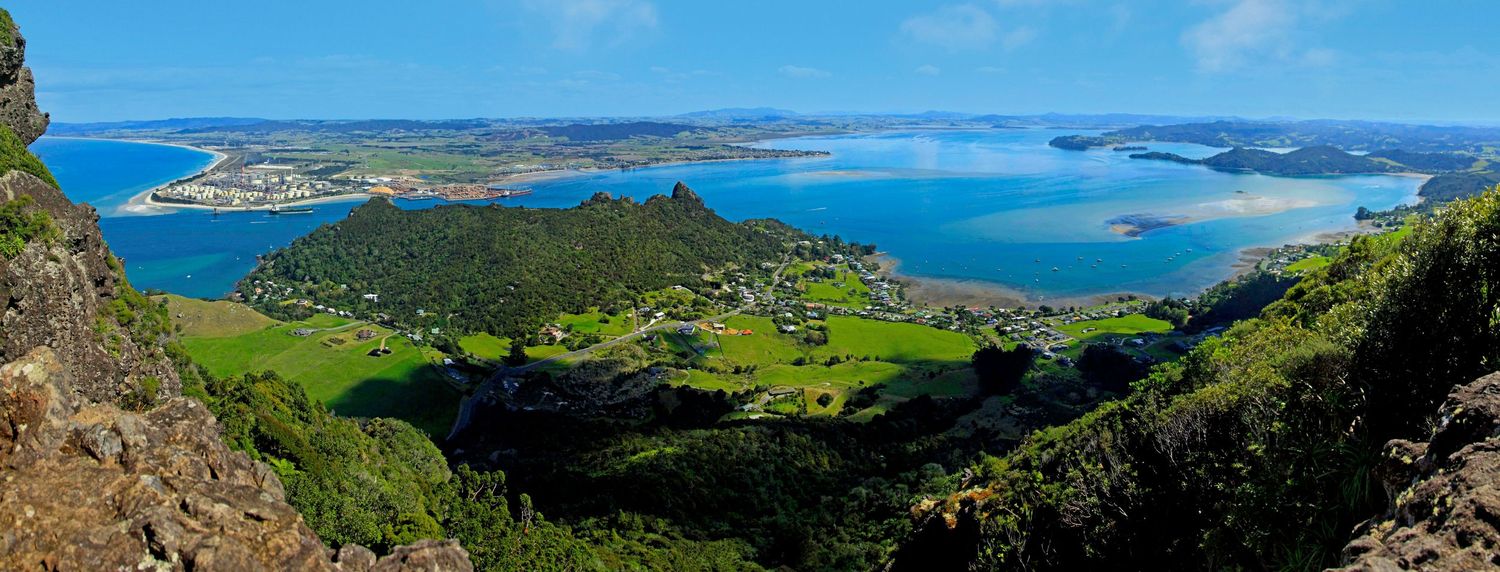 Panoramic aerial view of a coastal New Zealand town with lush green hills, blue harbor waters, and scenic bay landscape.
