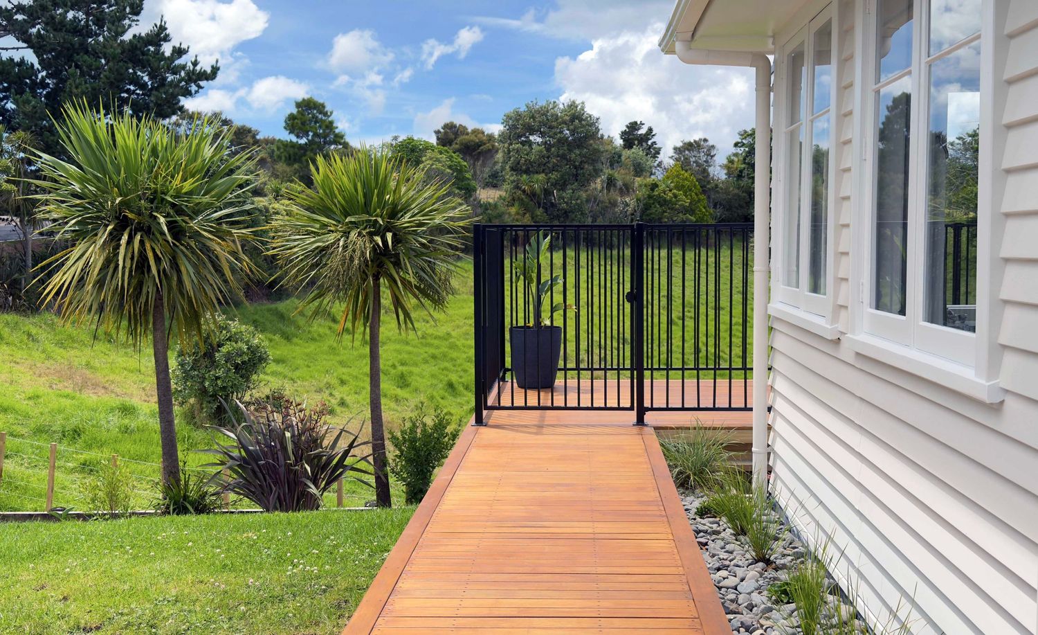 Wooden deck walkway leading to black metal gate surrounded by lush green lawn and palm trees beside white house.