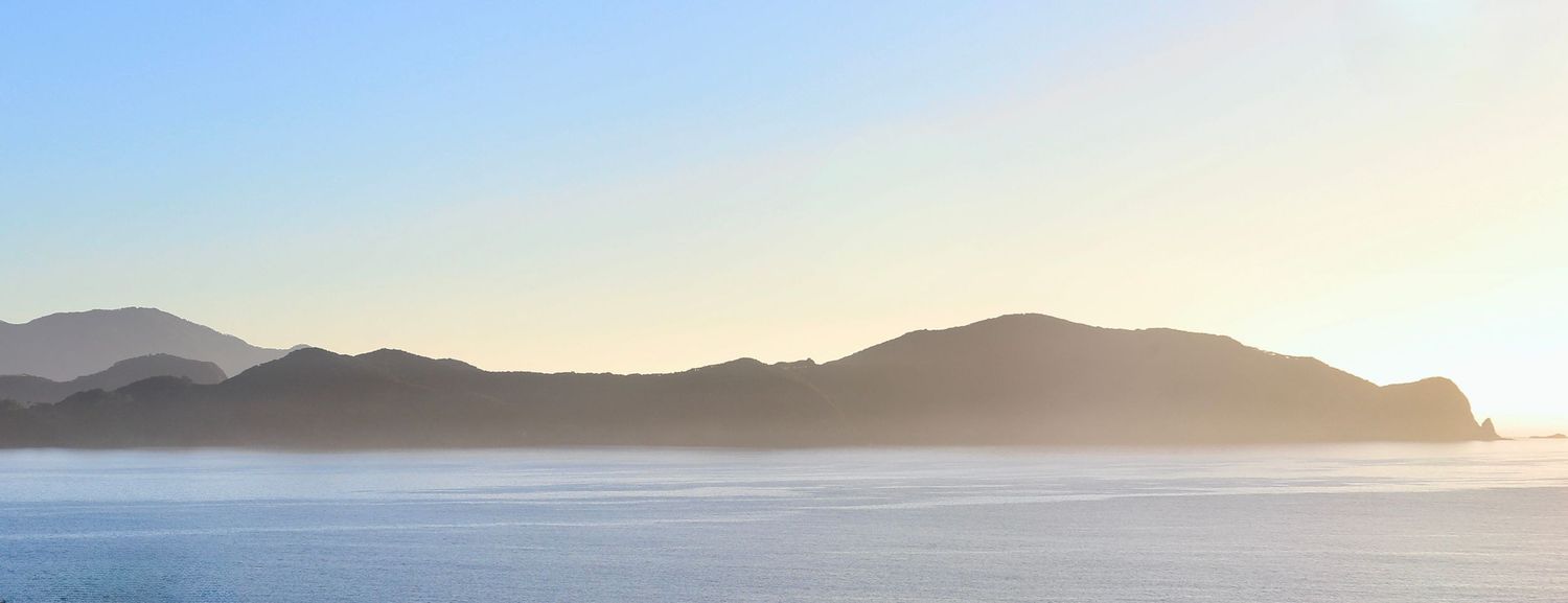 Serene coastal panorama with misty mountains silhouetted against a soft golden sky over calm blue ocean waters.