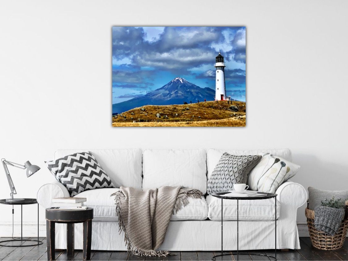 Canvas wall art of a white lighthouse with a volcanic mountain and dramatic blue cloudy sky in a bright living room.