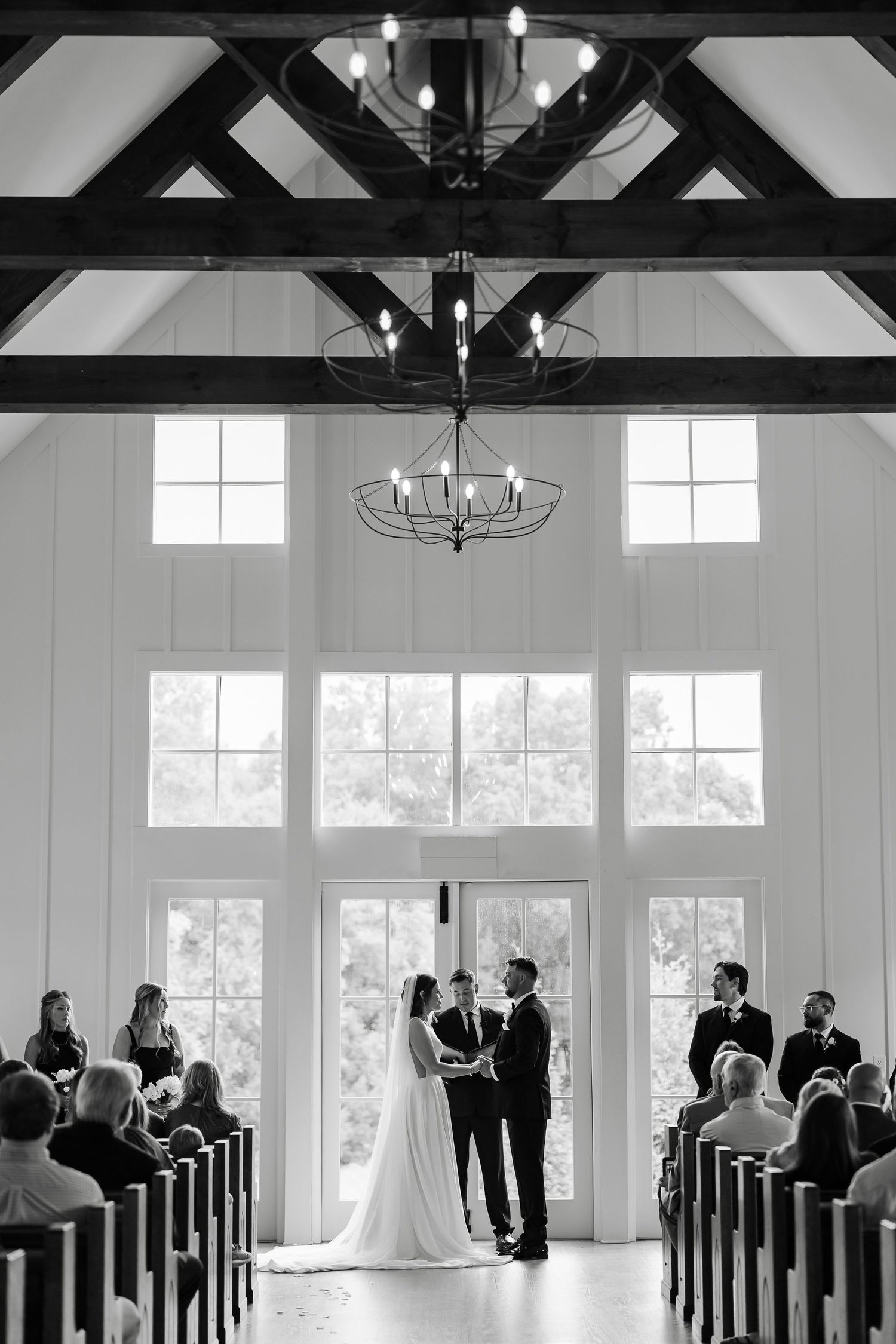 Black and white wedding ceremony photo inside a white chapel with cathedral ceilings, chandeliers, and large windows.At The Venue at Birchwo