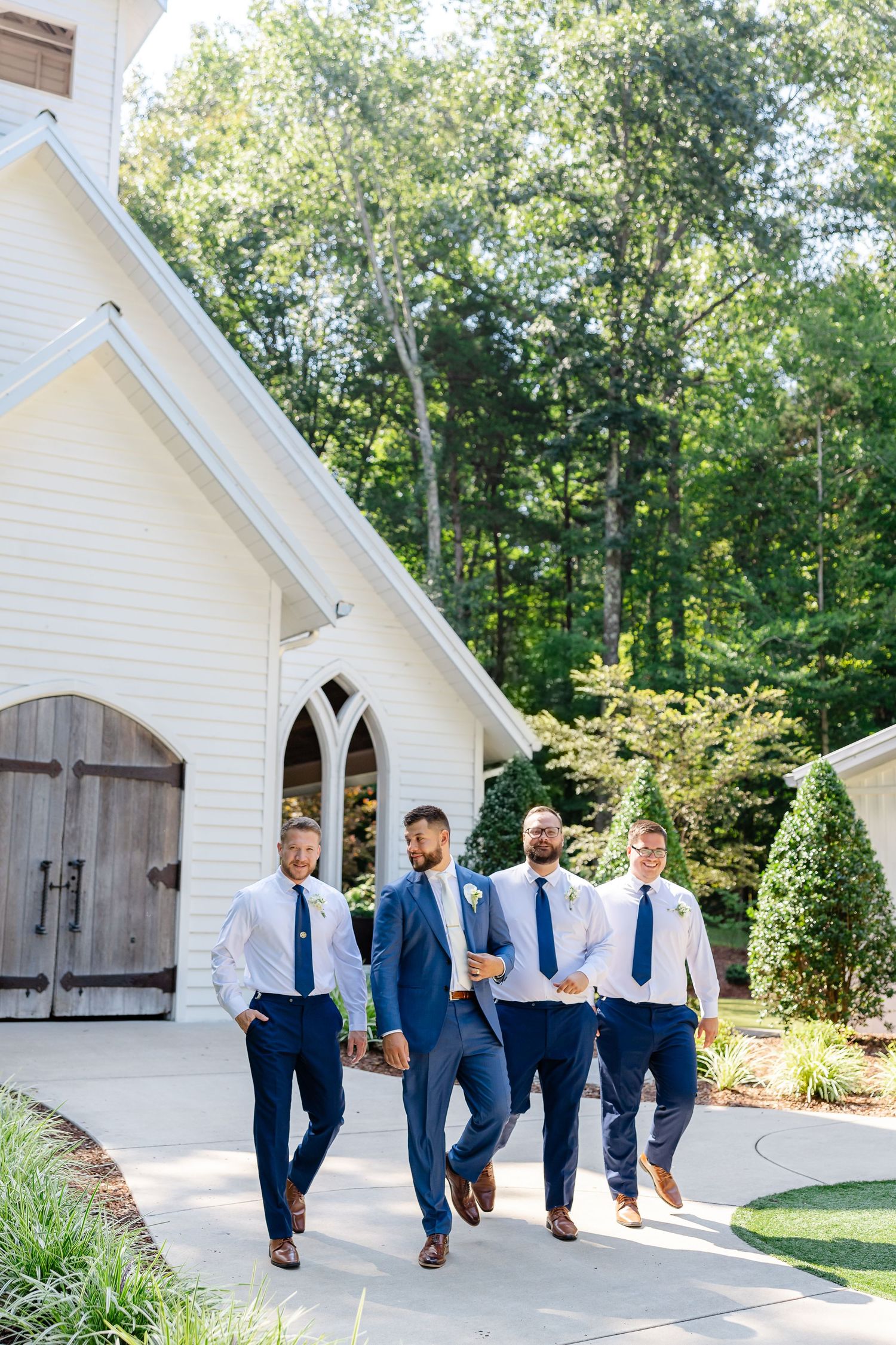 Four groomsmen in navy blue suits walk confidently outside a white chapel surrounded by lush green trees on a wedding day. At FireFly Lane