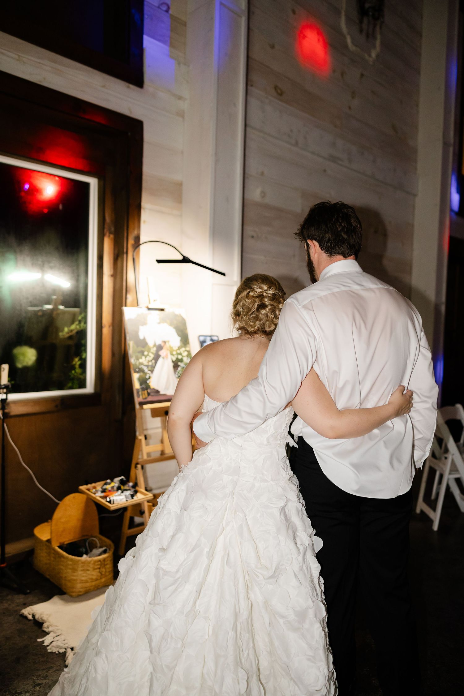 A bride in a white ruffled gown and groom in white shirt embrace from behind at their wedding reception.