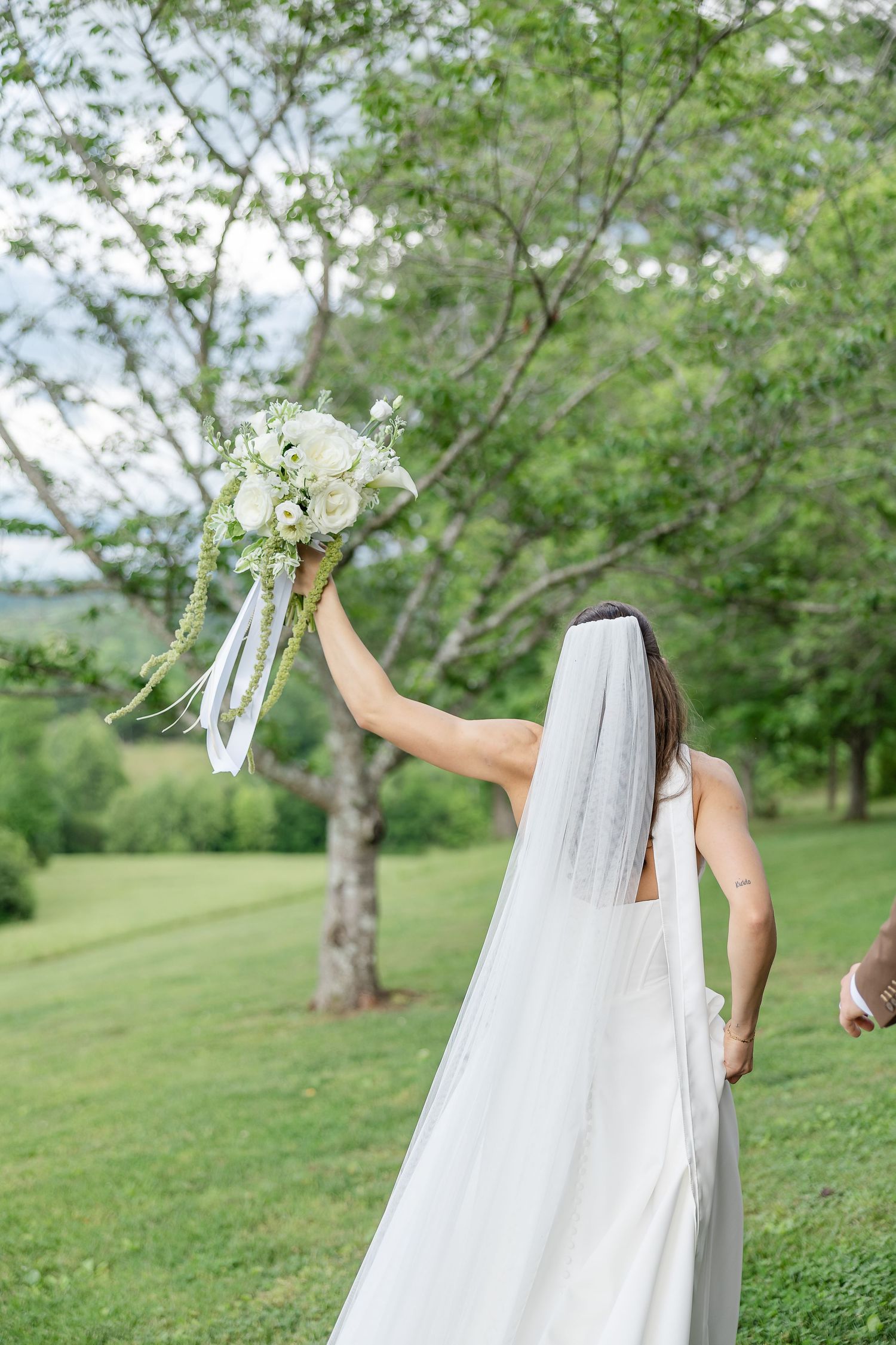 Bride in white gown and veil raises floral bouquet outdoors on lush green lawn surrounded by trees.