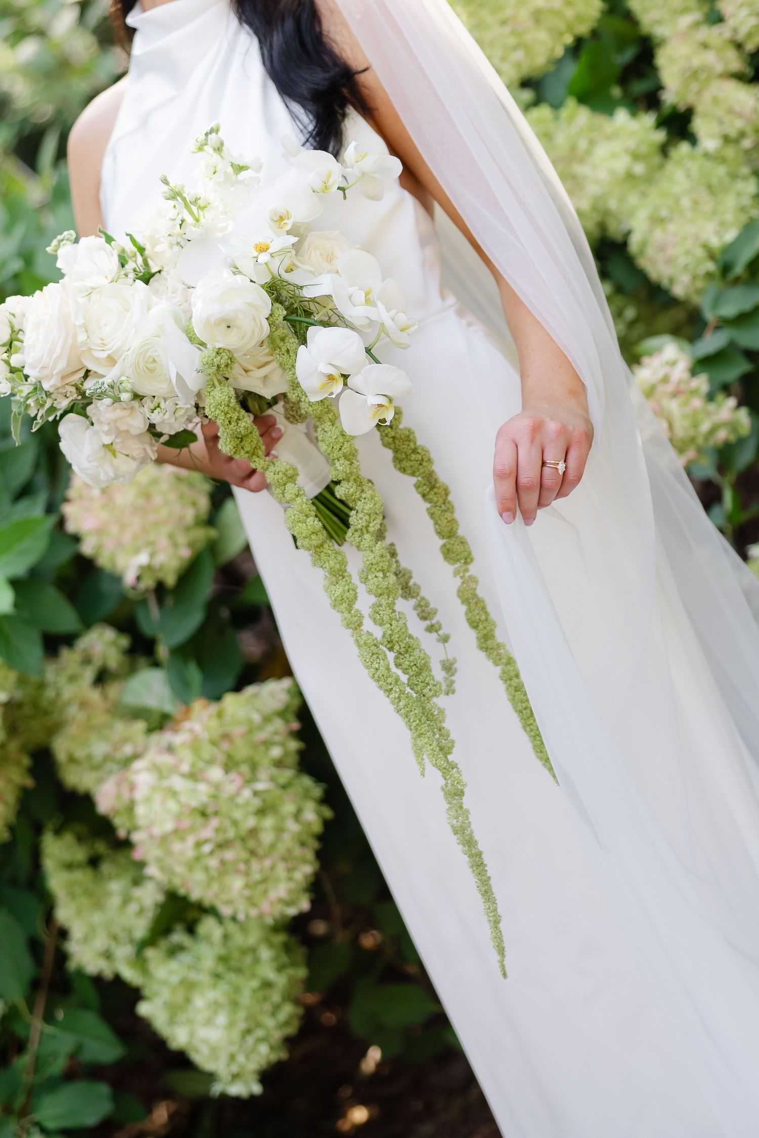 Bride in white gown holding cascading bouquet of white flowers and green amaranthus near hydrangea bushes. at firefly lane in Dickson TN