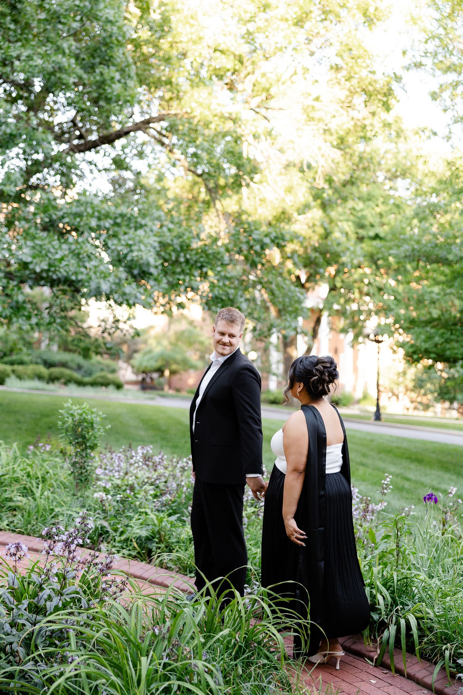 Couple in formal black attire walking hand in hand through a lush garden path, smiling back at camera.