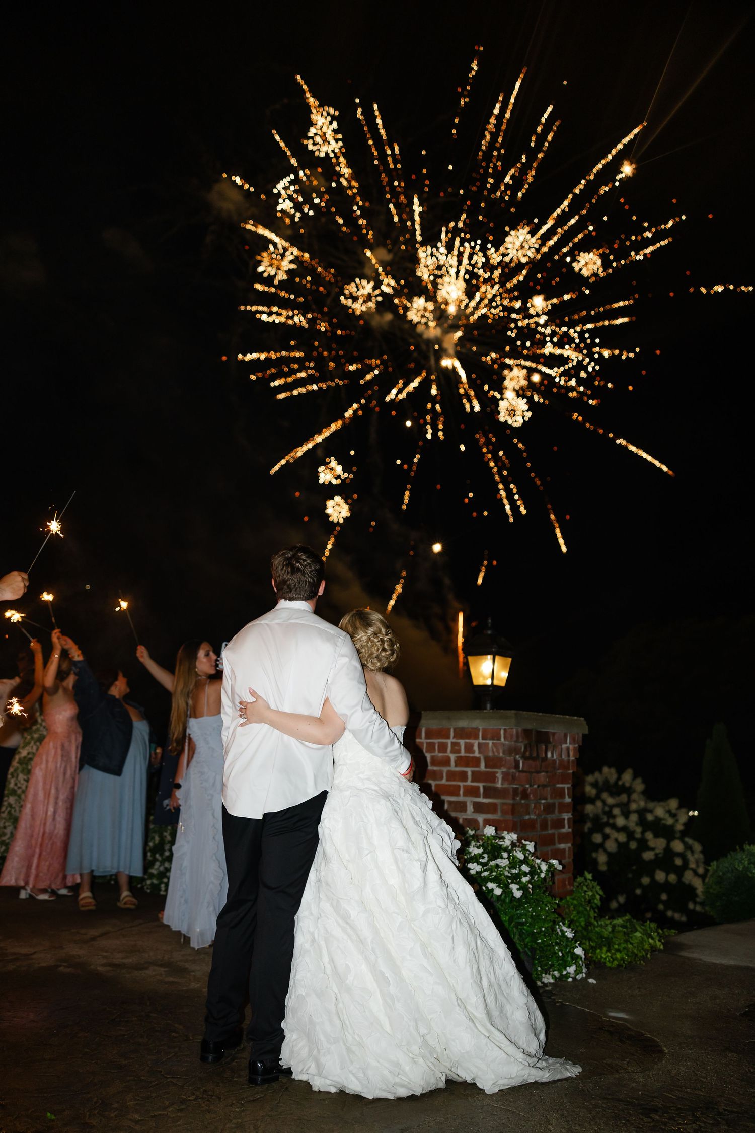 Bride and groom embrace watching golden fireworks burst in night sky, surrounded by guests holding sparklers at wedding reception.