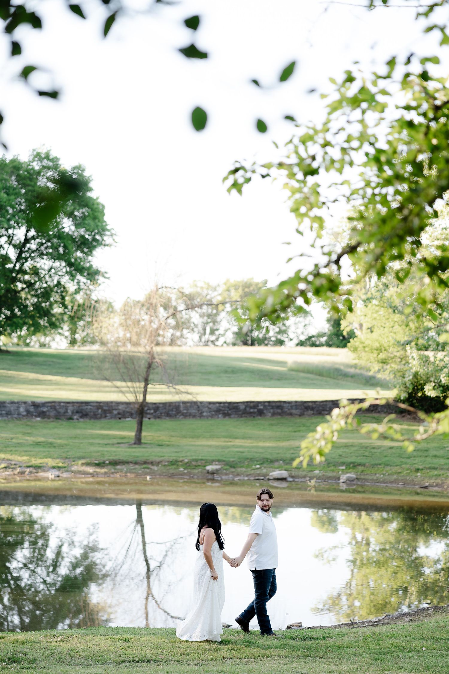 Bride and groom holding hands walking by a serene pond surrounded by lush green trees on a sunny day.