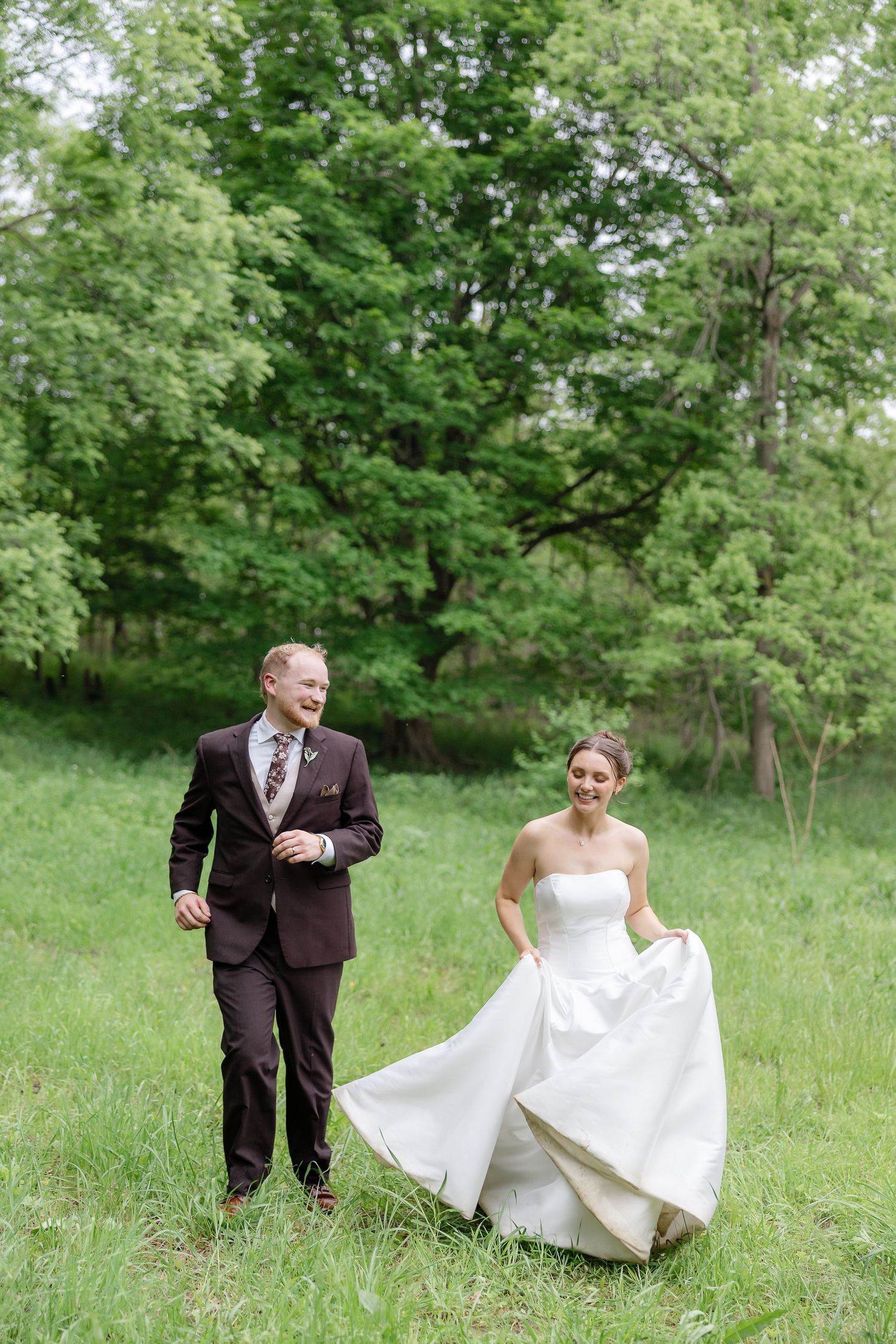 Bride and groom walking through lush green meadow, bride lifting white gown, groom in burgundy suit smiling.