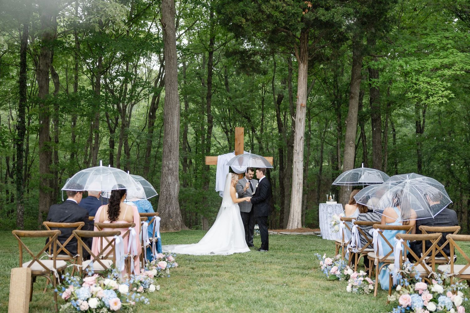 Outdoor rainy wedding ceremony in a lush forest with guests holding clear umbrellas as couple exchanges vows at a wooden cross.