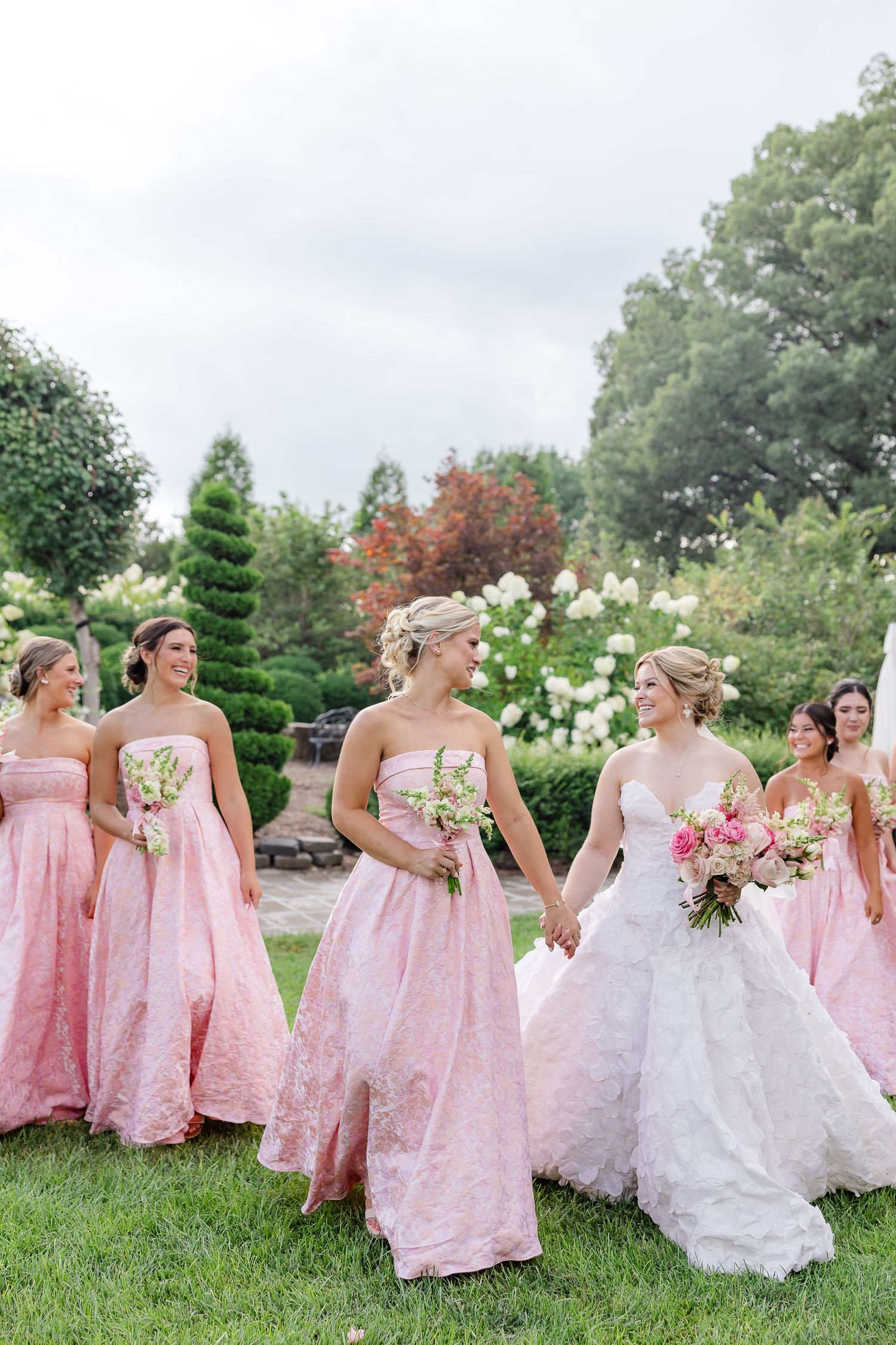 Bride in white gown holds hands with bridesmaid in pink strapless dress, walking through lush garden with bridal party.