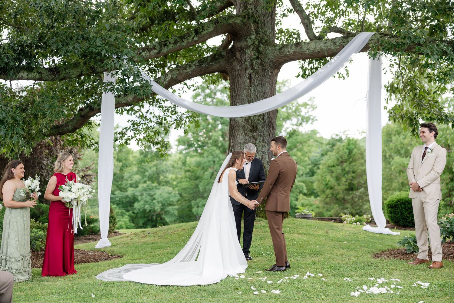 Outdoor wedding ceremony under a large tree with white draping, bride in white gown, groom in brown suit, officiant, and wedding party.