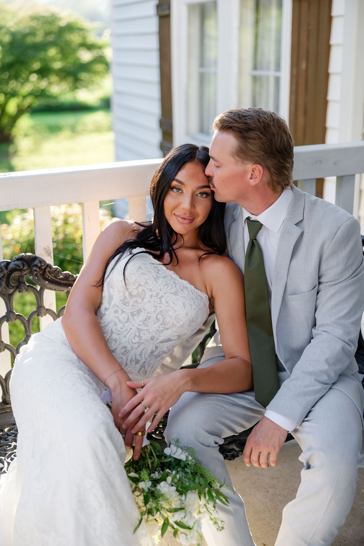 Bride and groom share a tender kiss on a porch bench, bride in lace gown, groom in gray suit with green tie. At Drakewood Farms in Nashville