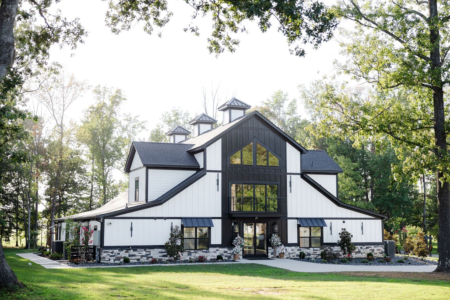 Modern white barn wedding venue with black roof, large windows, and lush green trees surrounding the property.