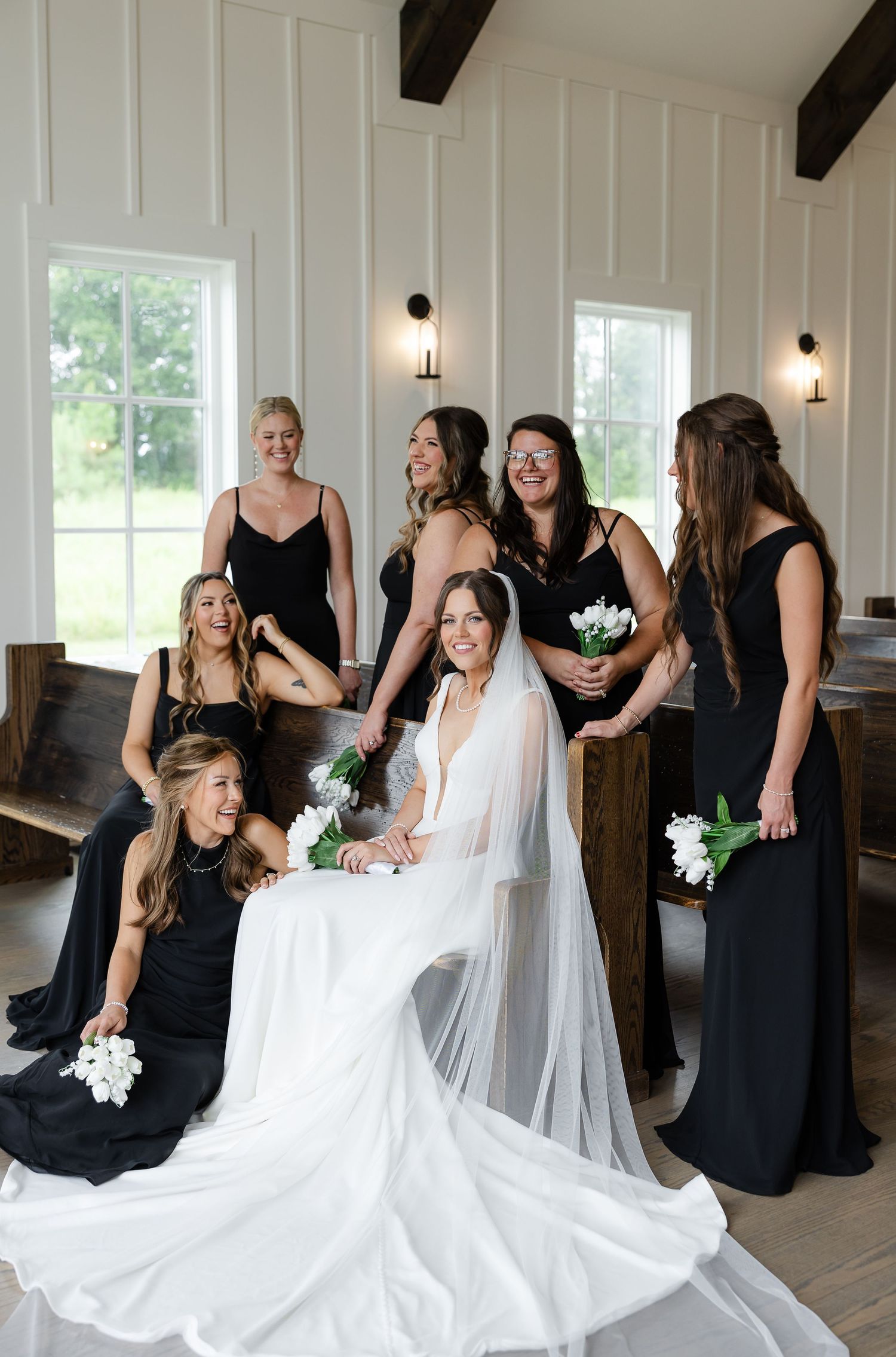Bride in white gown with bridesmaids in black dresses holding white tulips inside a white chapel. At The Venue at Birchwood