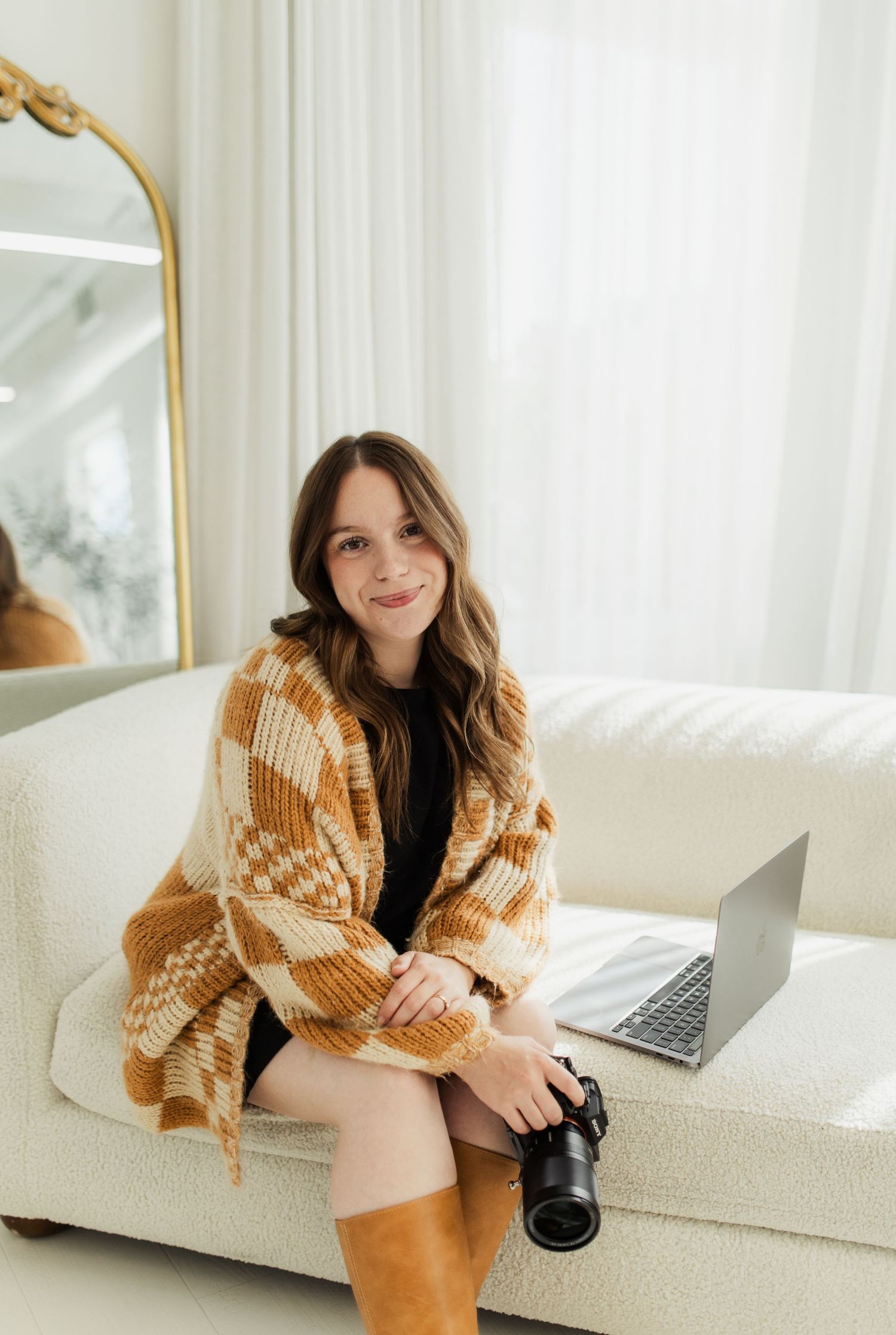 Young woman in plaid wrap shawl sits on white sofa holding camera with laptop, smiling in bright studio.