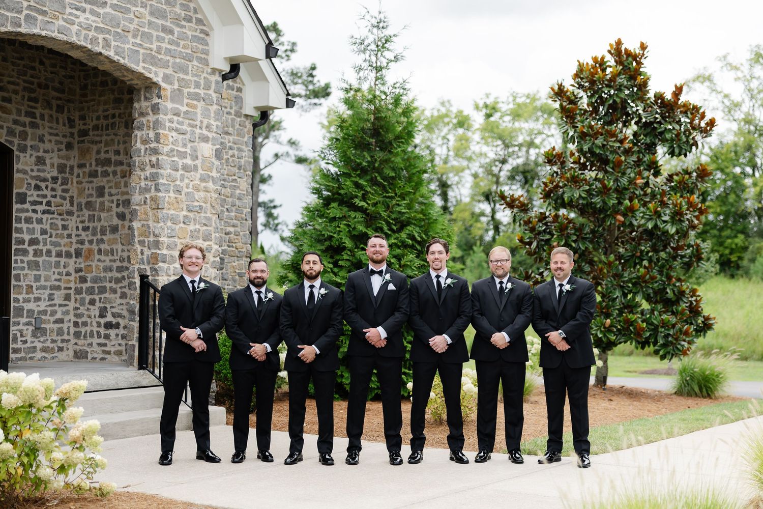 Groom and groomsmen in black suits posing outside a stone chapel surrounded by lush green trees on a wedding day. At The Venue at Birchwood