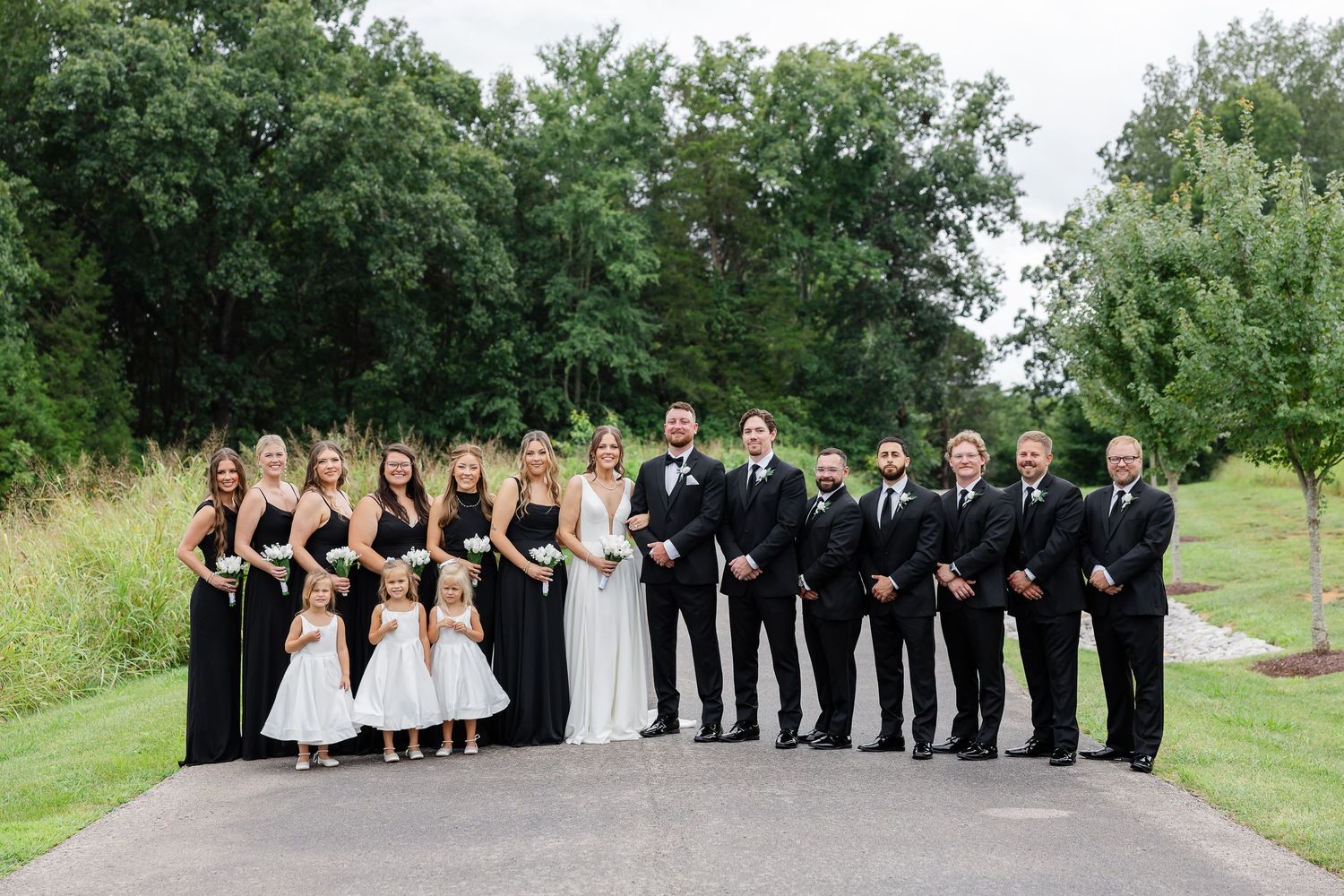 Wedding party in black attire with bride and groom posing on a tree-lined path outdoors.At The Venue at Birchwood