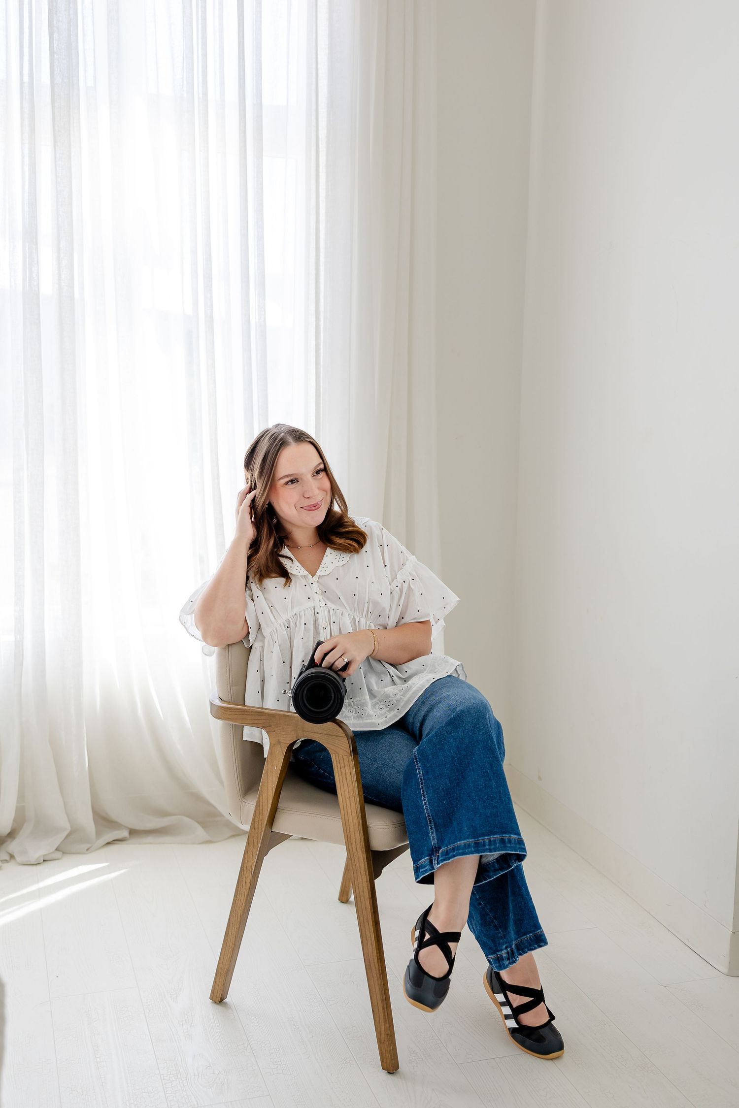 Woman in white blouse and jeans sits on wooden chair holding camera near sheer curtains in bright white studio.
