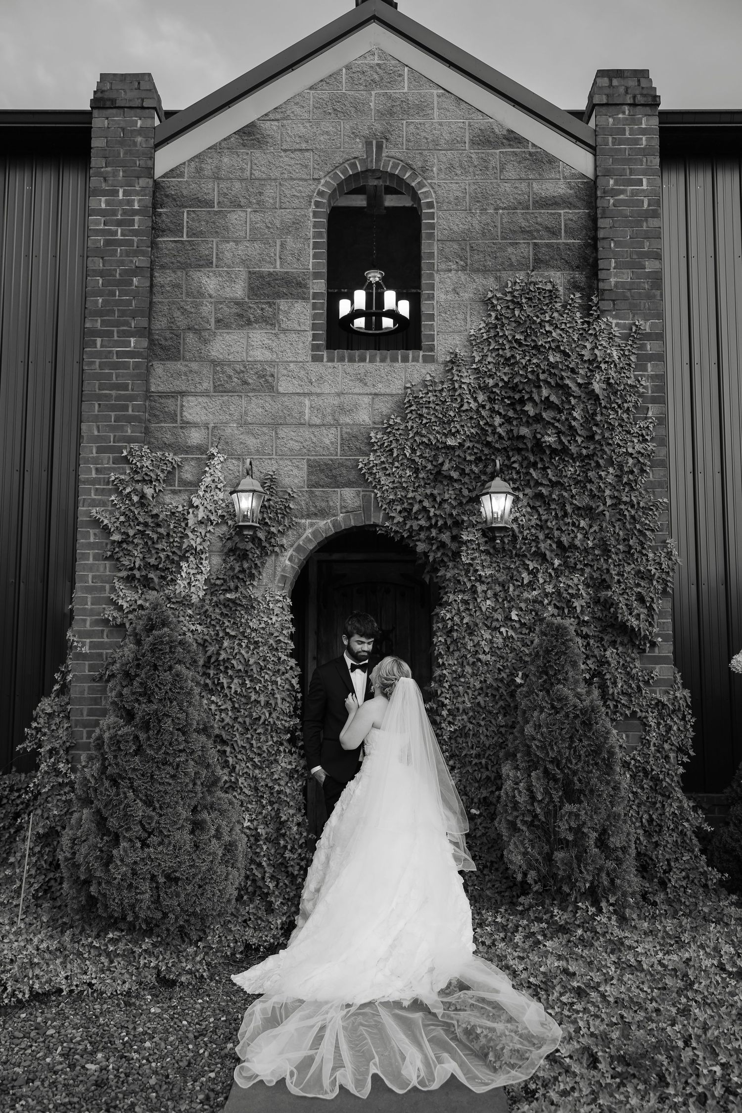Black and white photo of bride and groom posing at ivy-covered stone church entrance on their wedding day.